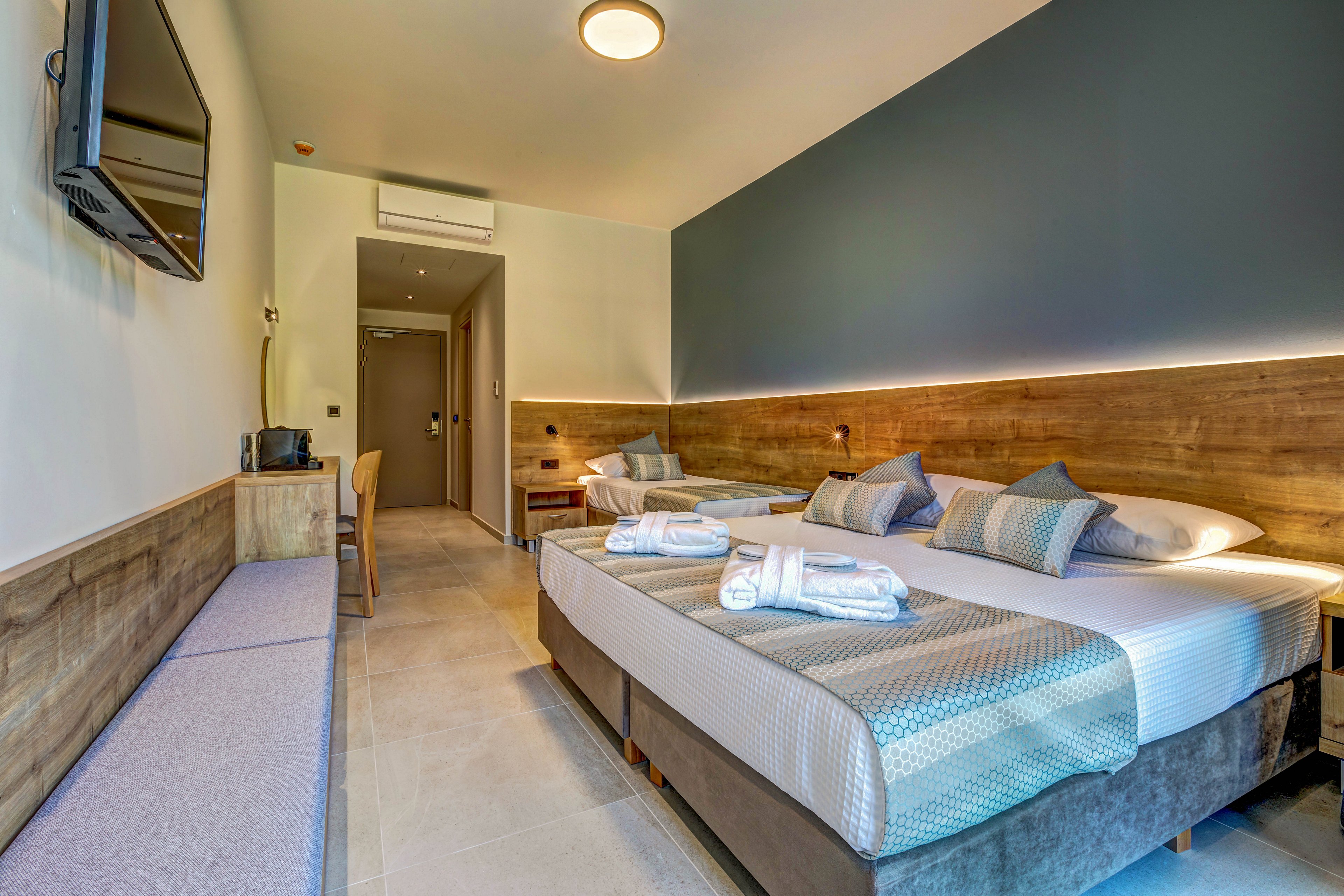 Contemporary hotel room interior featuring two neatly made beds with folded towels on top. The space includes a wall-mounted flat-screen TV, a long bench, and a small desk with a chair near the entrance. Warm wood accents, soft lighting, and blue and gray textiles create a calm, inviting atmosphere. The room appears clean, organized, and ready for arriving guests.