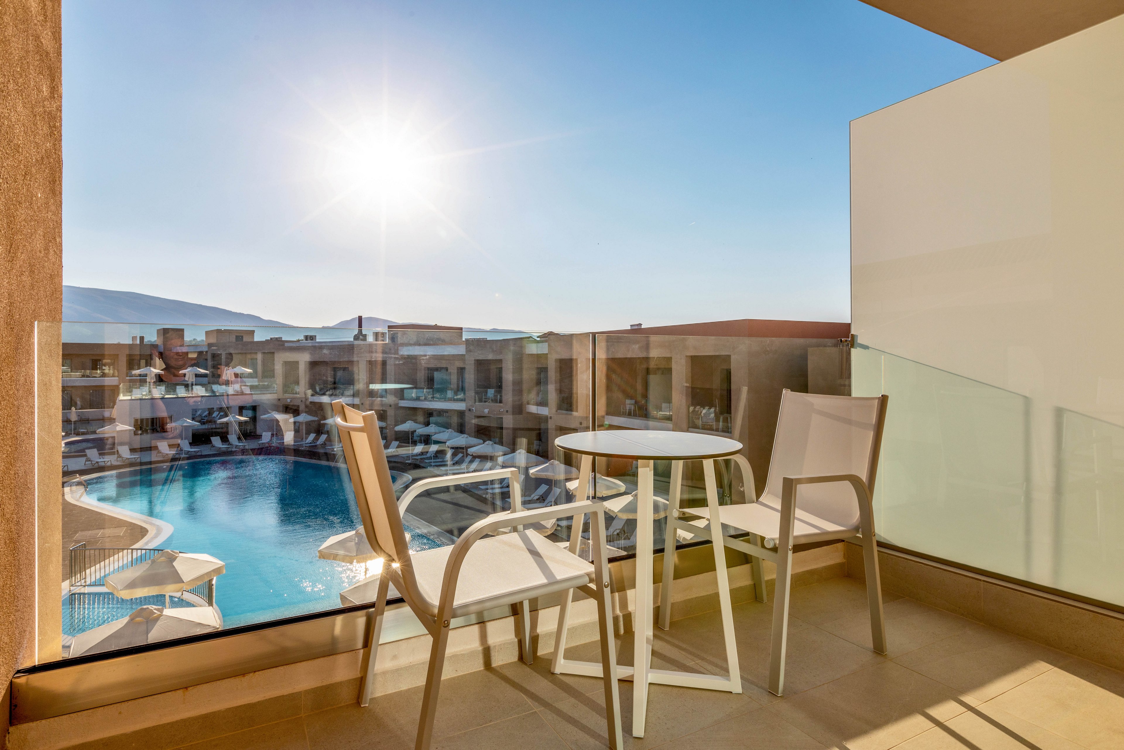 A modern hotel balcony features a small round table and two chairs facing a large swimming pool. The scene is bathed in bright sunlight with clear blue skies overhead. Glass railings frame the balcony, offering an unobstructed view of the resort buildings and pool area below. The overall atmosphere suggests a relaxing vacation setting in a warm, sunny destination.
