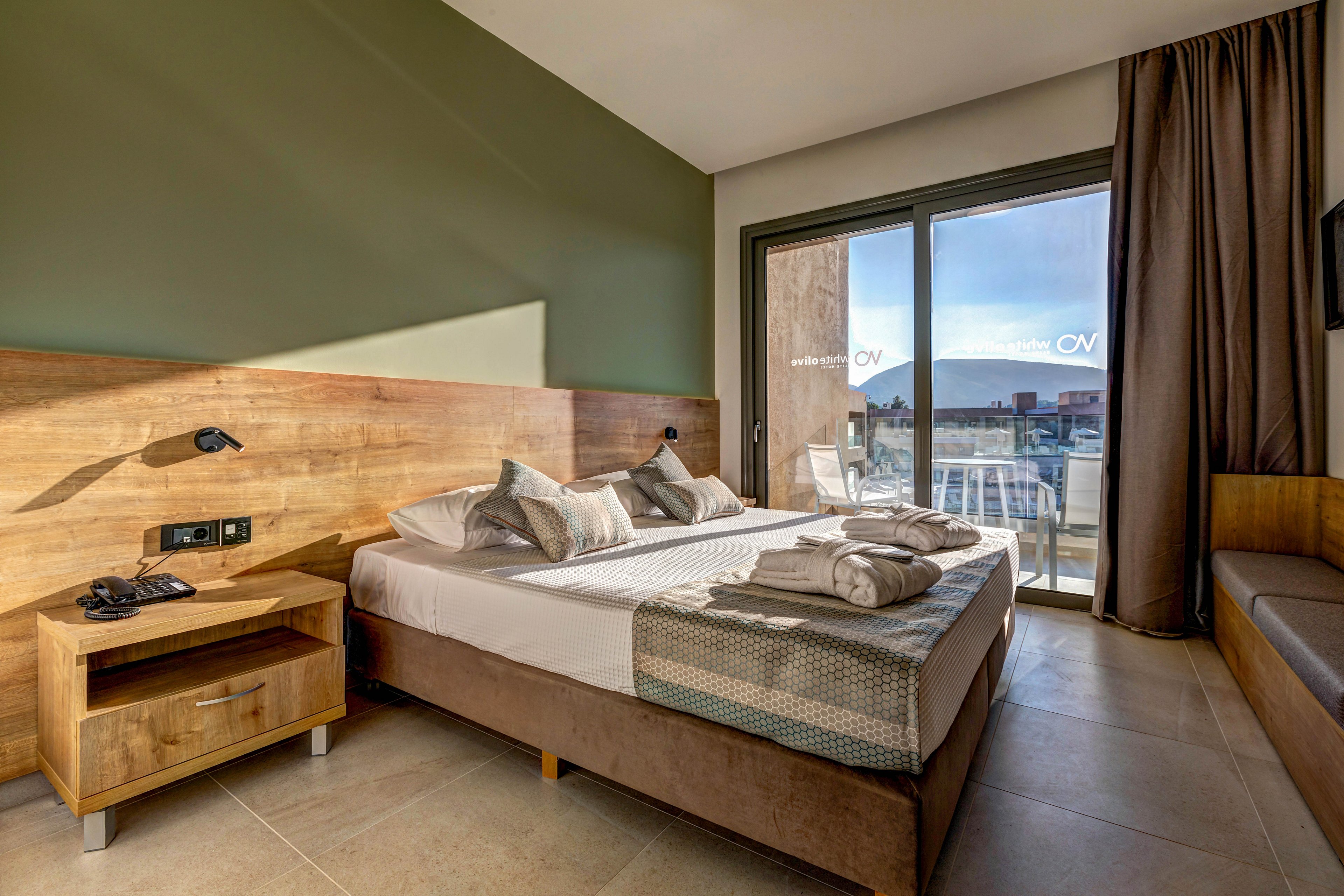 A contemporary hotel bedroom features a neatly made double bed with folded towels placed on top. The room is decorated with warm wooden furniture, including a bedside table with a telephone and reading lights. Large sliding glass doors open to a balcony with outdoor chairs and a view of surrounding buildings. Natural daylight fills the space, highlighting the neutral tones and clean, minimalist design.