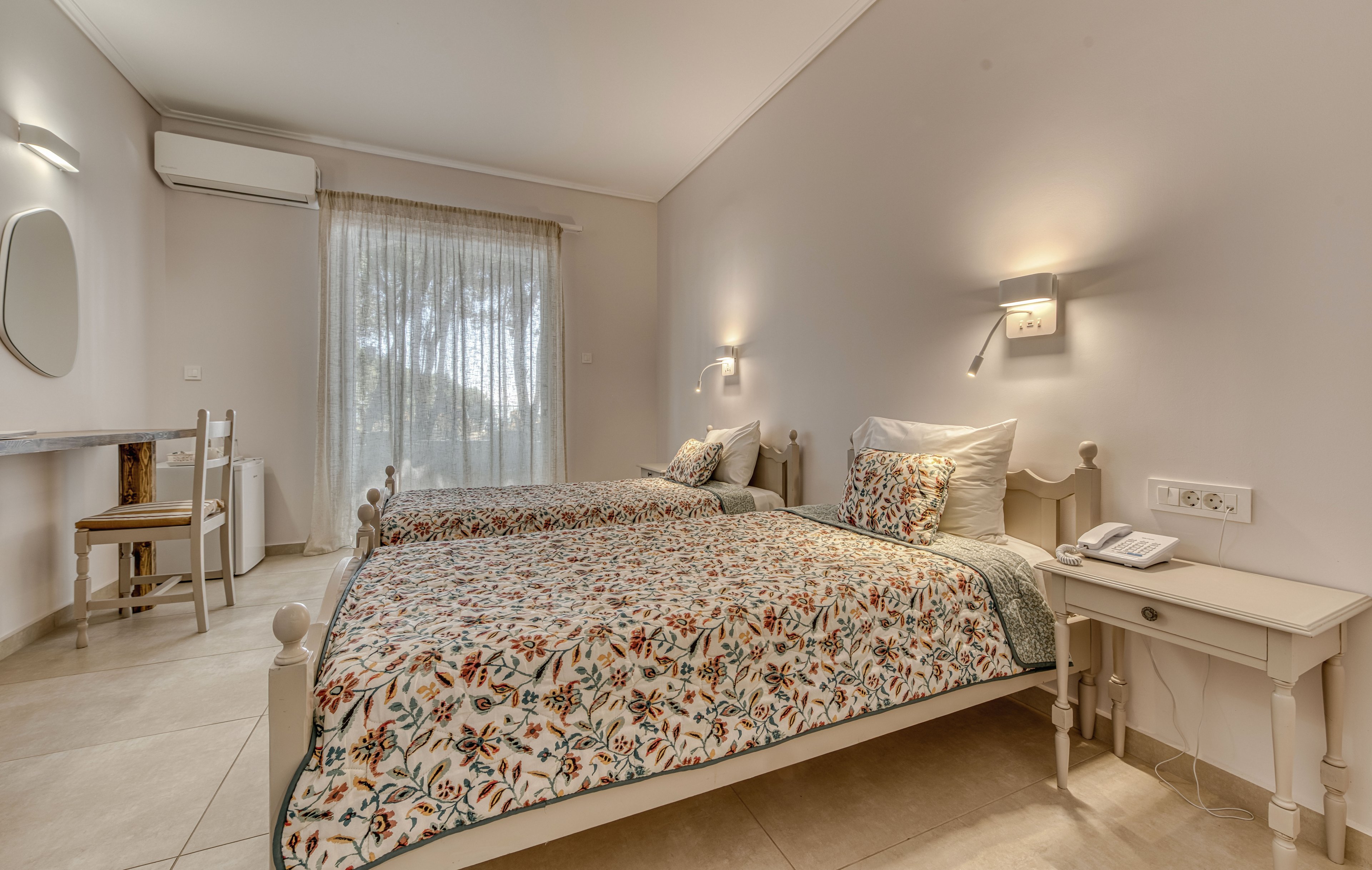 Bright, neutral-toned hotel room featuring two twin beds with floral patterned bedspreads. The space includes a small bedside table with a telephone and a wall-mounted reading light above each bed. A desk with a chair and a round mirror sits against the opposite wall, while sheer curtains cover a large window or glass door. The overall atmosphere is clean, simple, and inviting, suitable for guests seeking a comfortable stay.