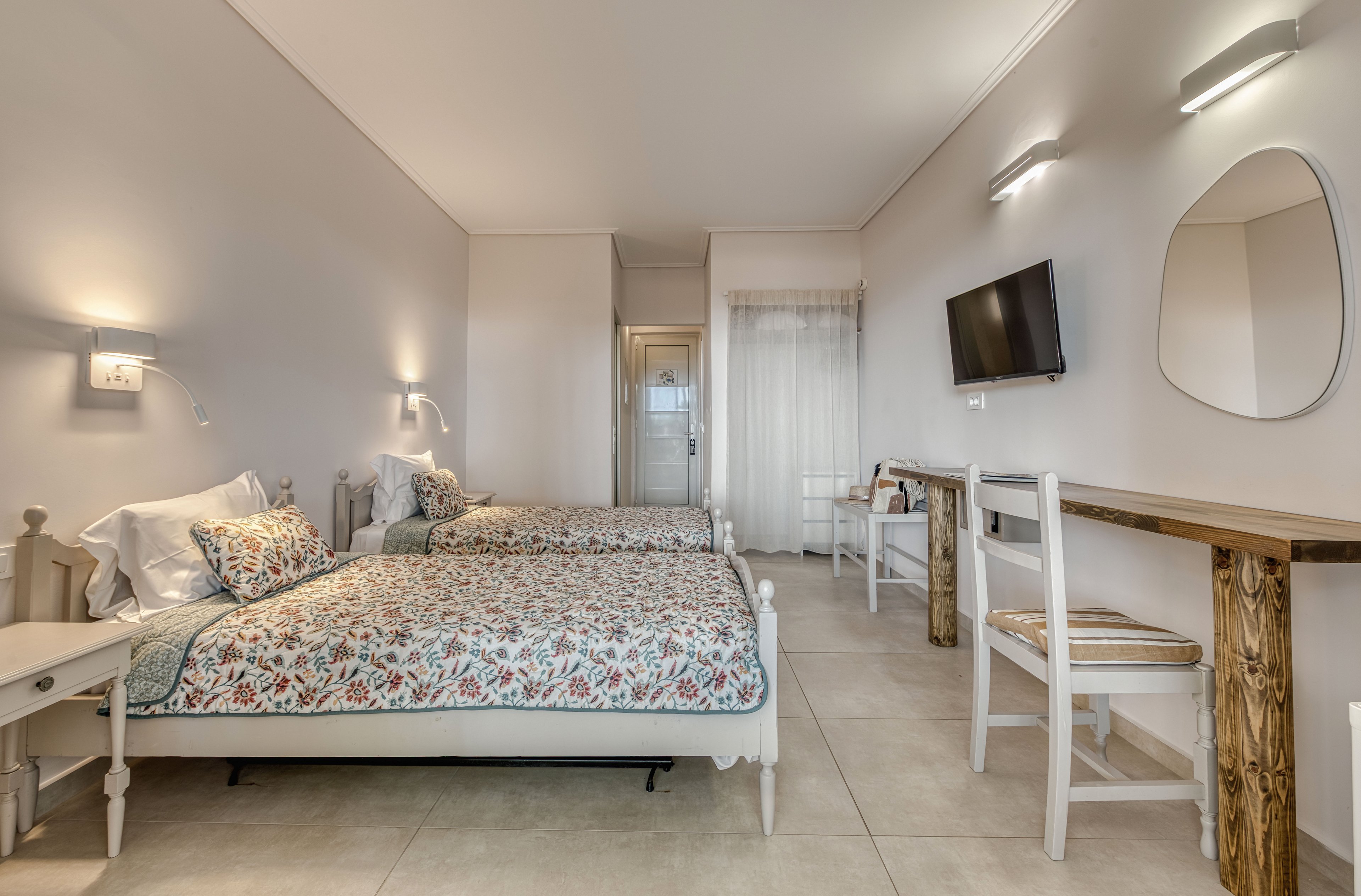 A cozy hotel room features two twin beds with floral patterned bedding and white wooden frames. The space is bright and minimal, with light-colored tile flooring and soft neutral walls. A wall-mounted flat-screen television faces the beds, accompanied by a long wooden console table and simple white chairs. Soft lighting and sheer curtains create an inviting, relaxed atmosphere.