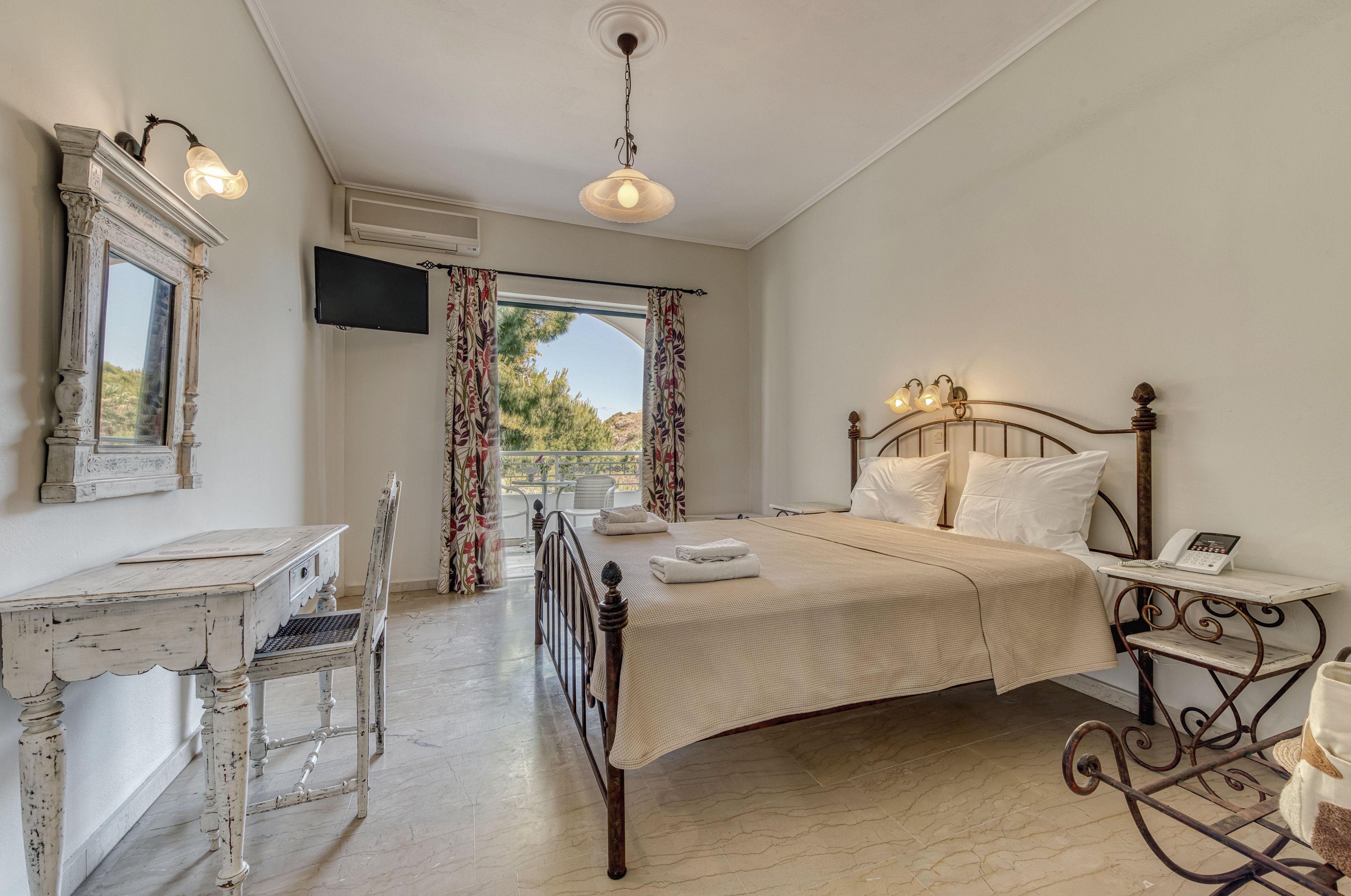 Cozy double bedroom with a metal bed frame is styled in a vintage aesthetic with distressed white furniture. The room features a small desk and chair, wall-mounted television, and bedside table with lamp. Floral curtains frame a glass door leading to a balcony with greenery and trees visible outside. Neutral tones and soft lighting create a calm, inviting atmosphere suitable for a relaxing stay.