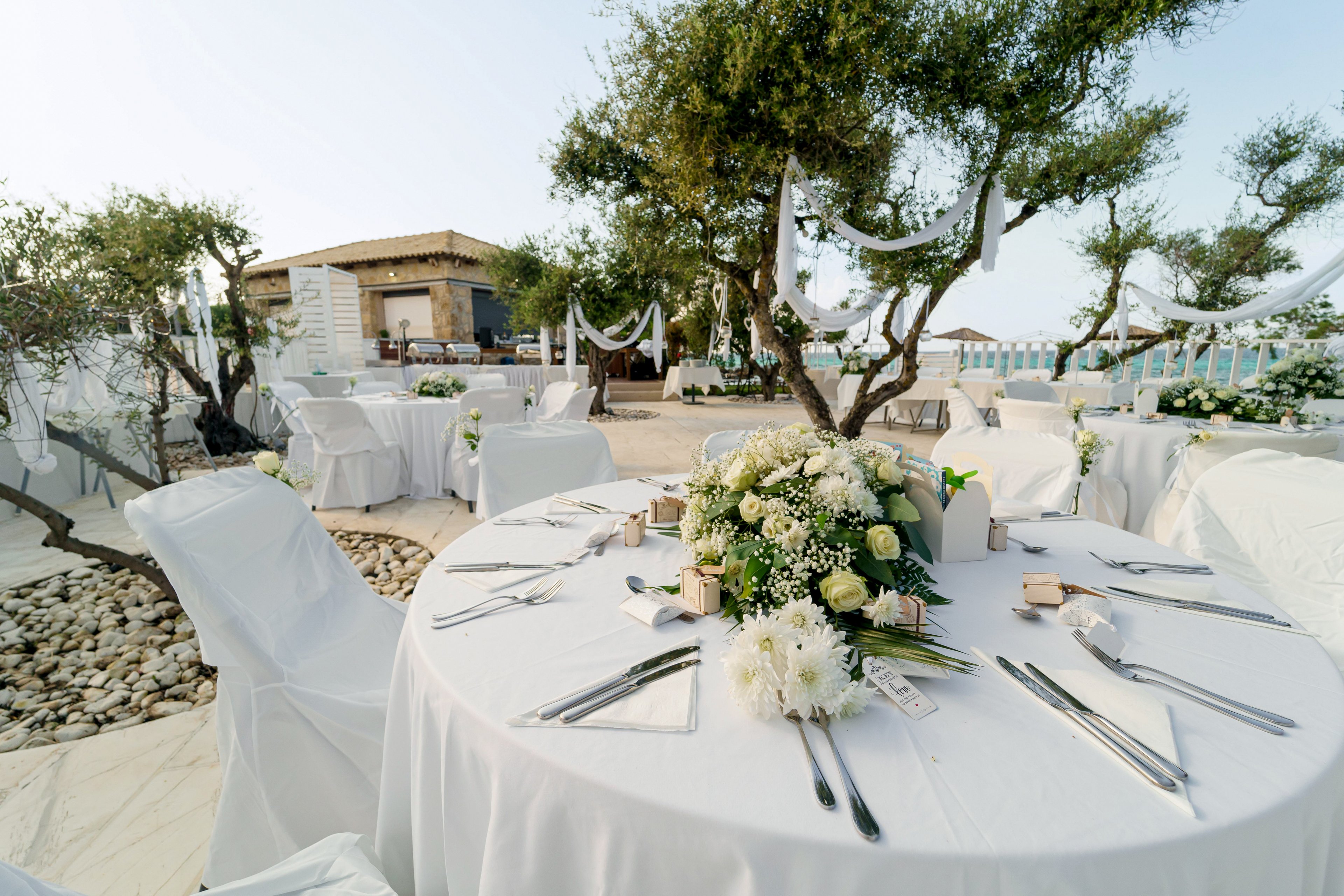 Round tables with white linens are set for an elegant outdoor wedding reception. Floral centerpieces with white and green flowers decorate each table, surrounded by neatly arranged cutlery and glassware. The venue is located near the sea, with a view of the water and a rustic building in the background. Trees wrapped with white fabric and hanging decorations add a romantic, festive atmosphere to the scene.