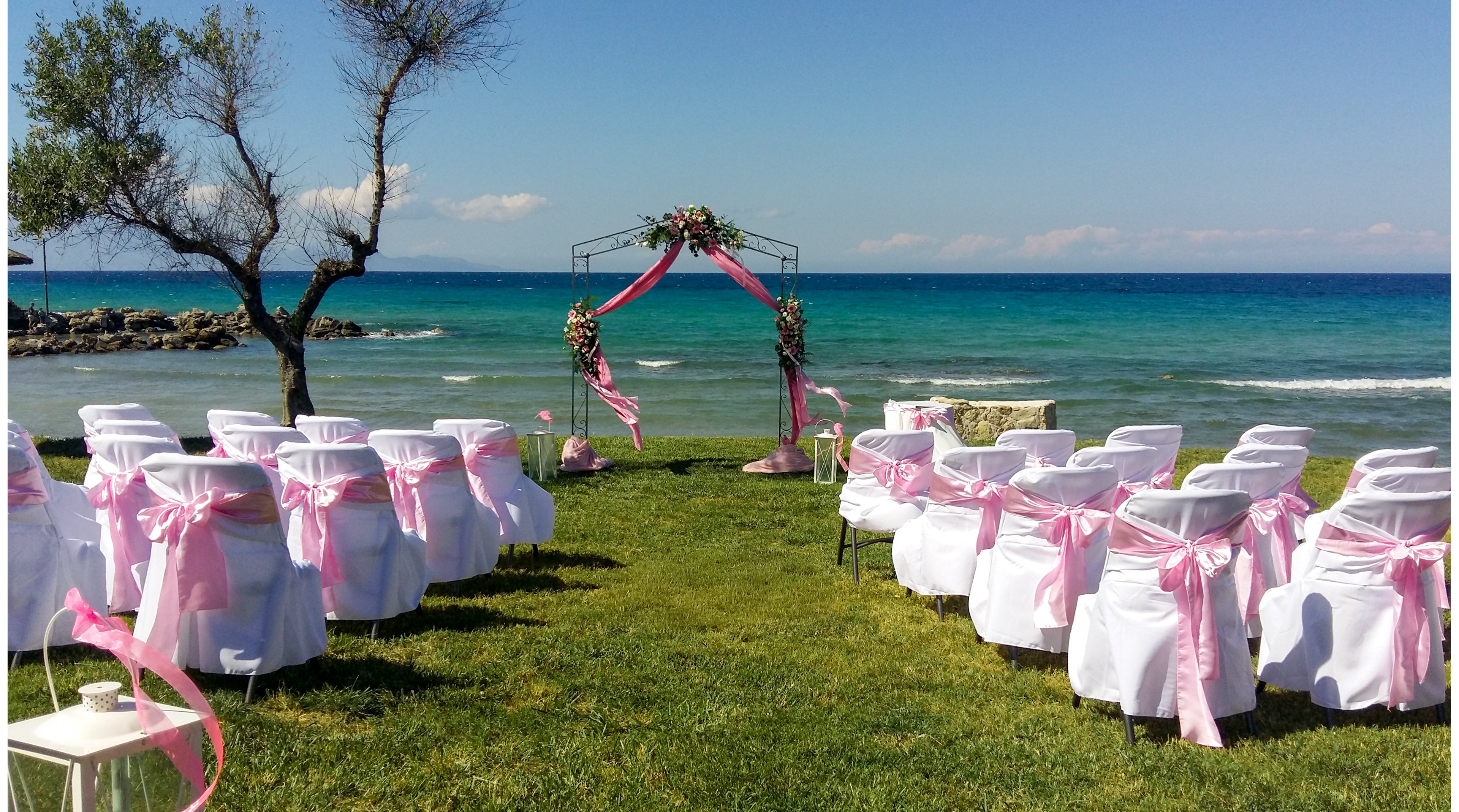 An outdoor wedding ceremony setup is arranged on a grassy lawn beside the sea. White chairs decorated with pink ribbons face a floral archway overlooking the blue water. A few small trees and a clear horizon line frame the scenic coastal backdrop. The scene feels bright, romantic, and ready for guests to arrive.