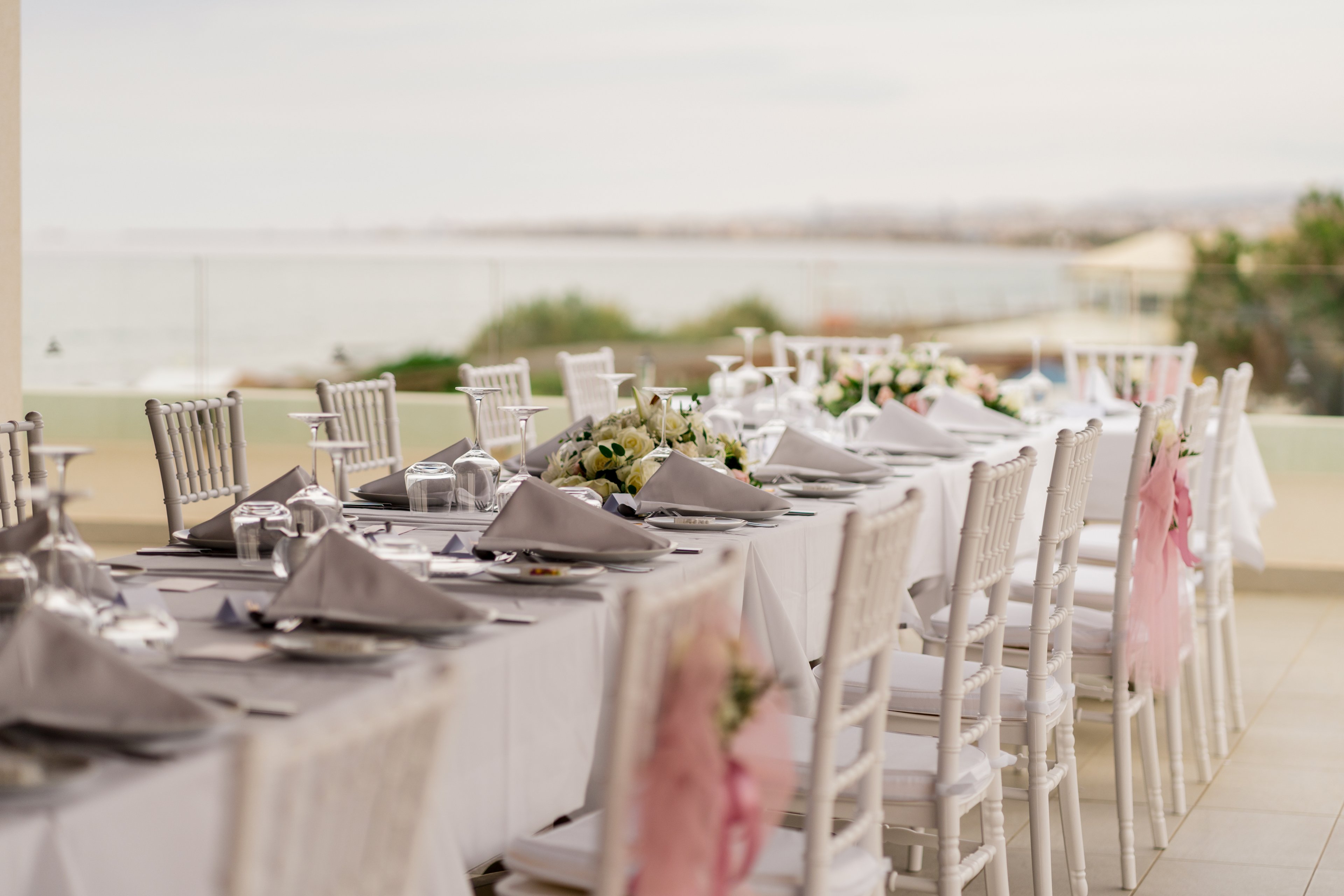 A long, elegantly decorated table is set for a formal event on an outdoor terrace. White chairs and tablecloths create a clean, sophisticated look, complemented by neatly folded gray napkins and polished glassware. Floral centerpieces with white and green flowers run along the middle of the table. The background shows a soft, out-of-focus view of water and distant shoreline, adding a serene coastal atmosphere.
