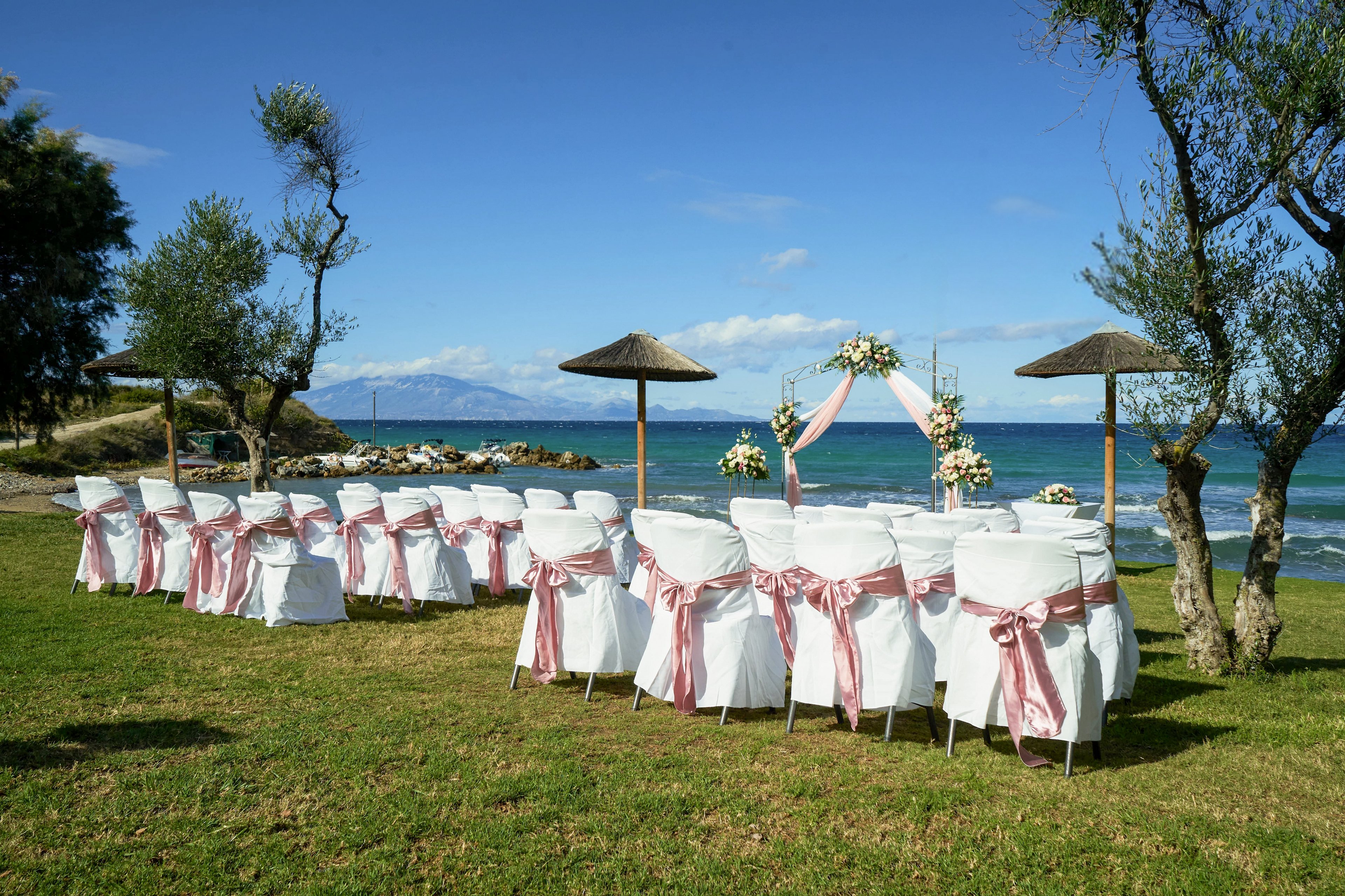 Rows of white chairs with pink ribbons are arranged on a grassy lawn facing a floral wedding arch by the sea. The ceremony setup overlooks calm blue water and distant mountains under a clear sky. Trees and thatched parasols frame the scene, adding a natural, relaxed atmosphere. The overall setting suggests a romantic beachside wedding ready for guests to arrive.