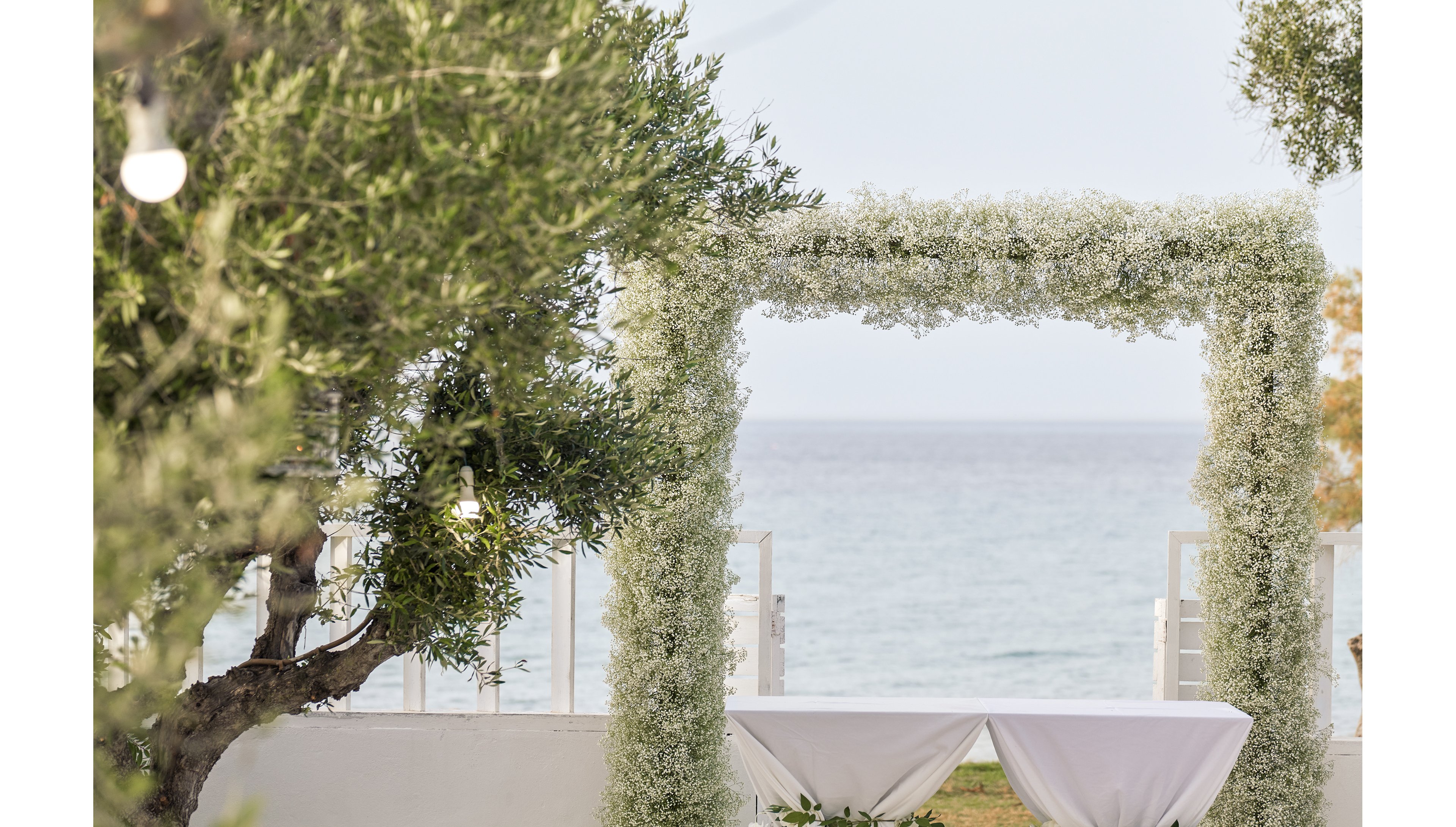 A square floral arch covered in delicate white flowers stands in an outdoor setting near the sea. Two white-covered tables are placed beneath the arch, suggesting preparations for a ceremony or reception. Olive trees and hanging lights frame the scene, adding a natural and romantic touch. The calm water and soft daylight create a serene coastal atmosphere.