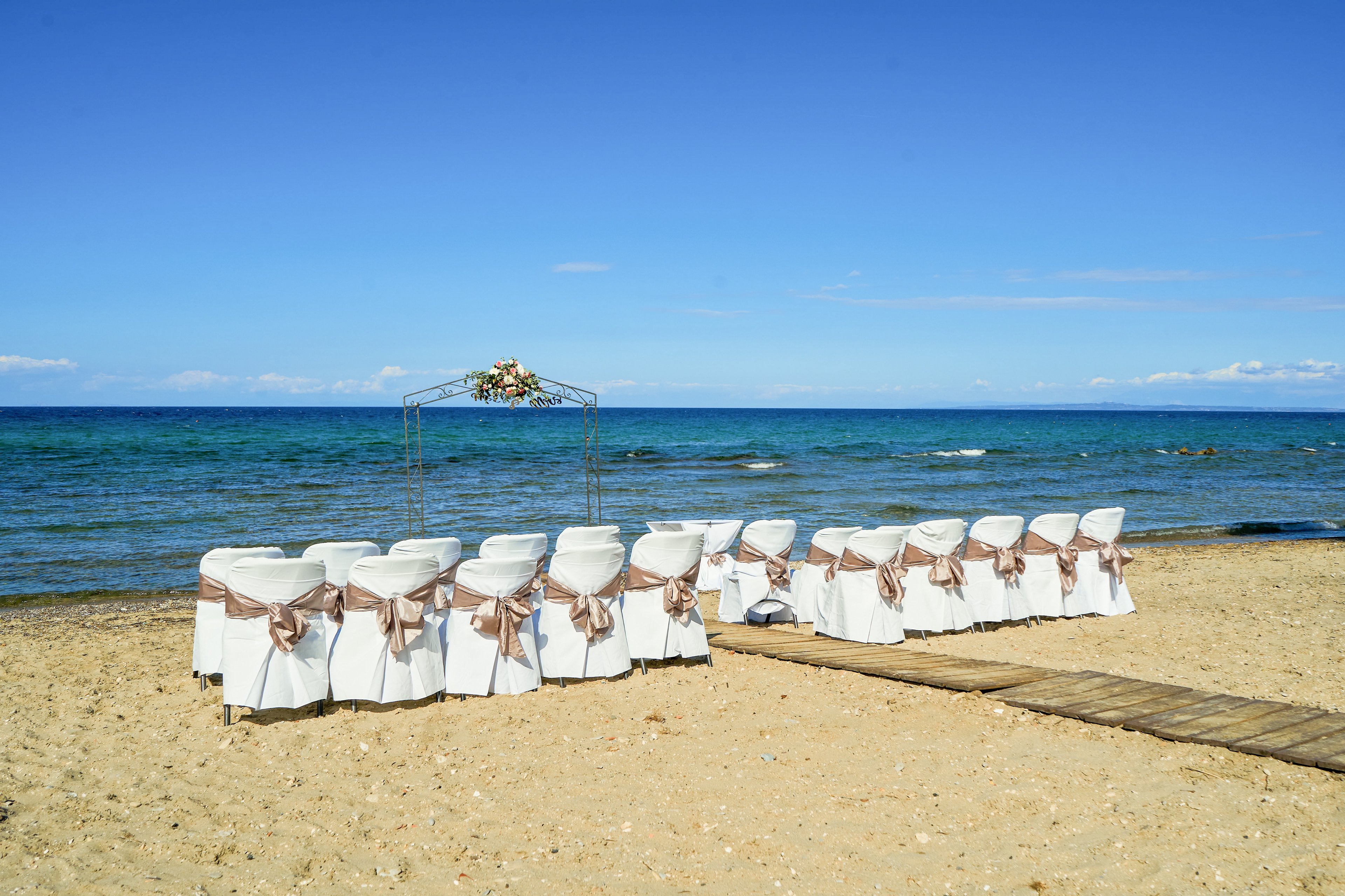 Rows of white-covered chairs with satin bows are arranged on a sandy beach facing the ocean. A simple floral arch stands at the front, framing the view of the blue water and horizon. The scene is calm and bright under a clear sky, suggesting a romantic seaside wedding ceremony. No guests are present yet, emphasizing the peaceful, anticipatory atmosphere.
