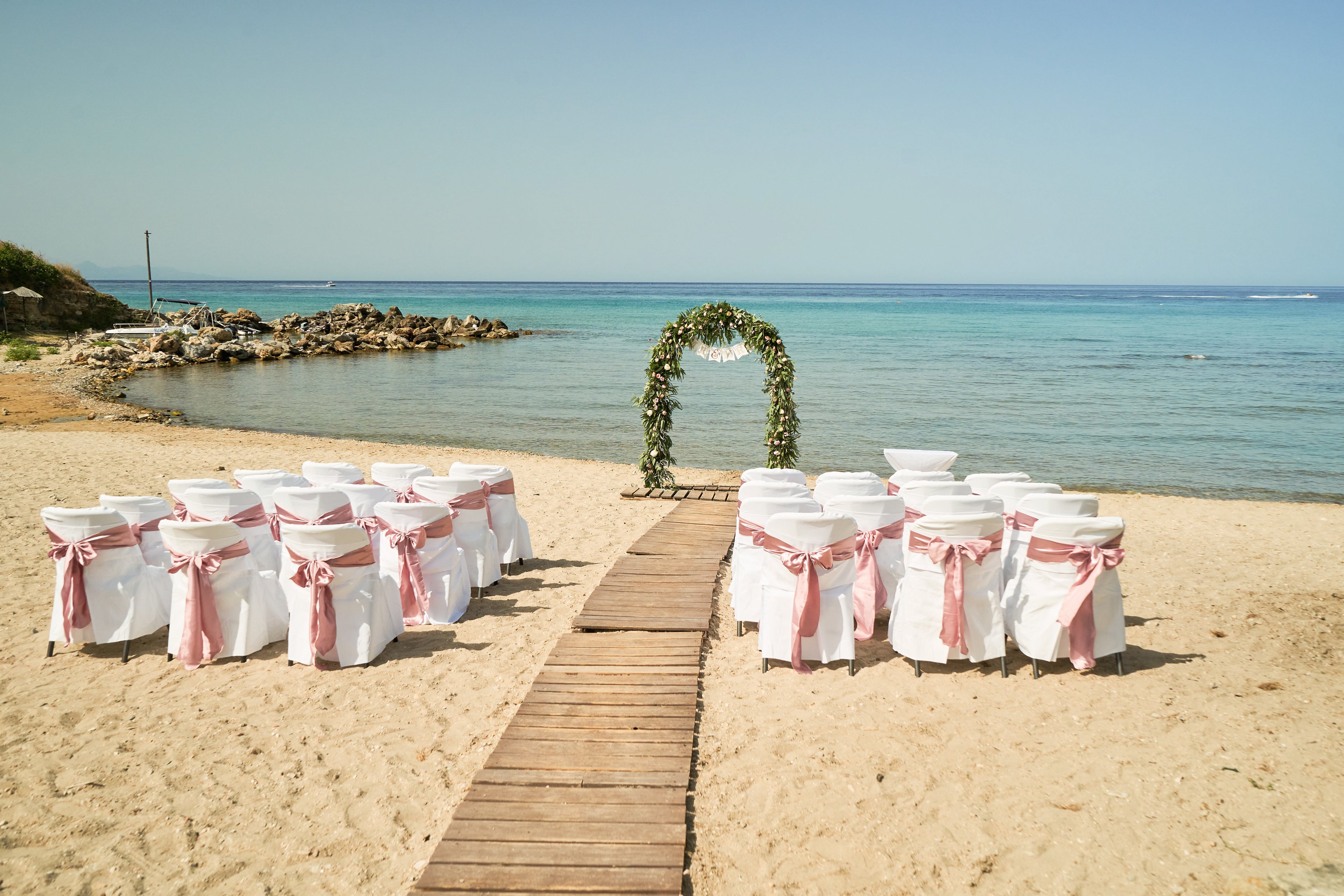 A wedding ceremony area is arranged on a sandy beach facing the calm sea. White chairs with pink sashes are lined up on both sides of a wooden walkway. At the end of the path stands a floral arch decorated with greenery and white flowers. The clear sky and tranquil water create a serene, romantic atmosphere for an outdoor seaside wedding.
