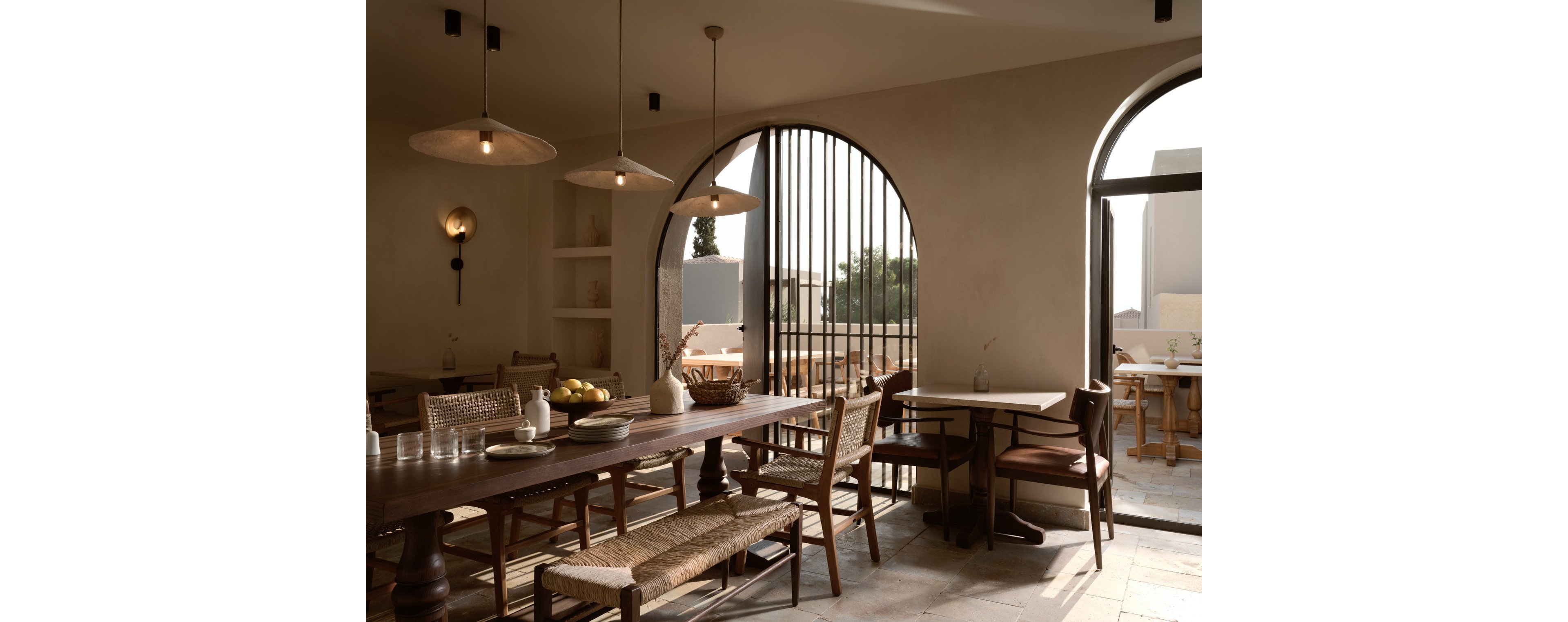 A warm, minimalist cafe interior features wooden tables and woven chairs arranged in a spacious dining area. Large arched windows and doors allow natural light to pour in, casting soft shadows across the stone floor. Simple pendant lights and wall sconces add subtle illumination to the neutral-toned space. The overall atmosphere feels calm, airy, and inviting, with a focus on natural materials and clean lines.
