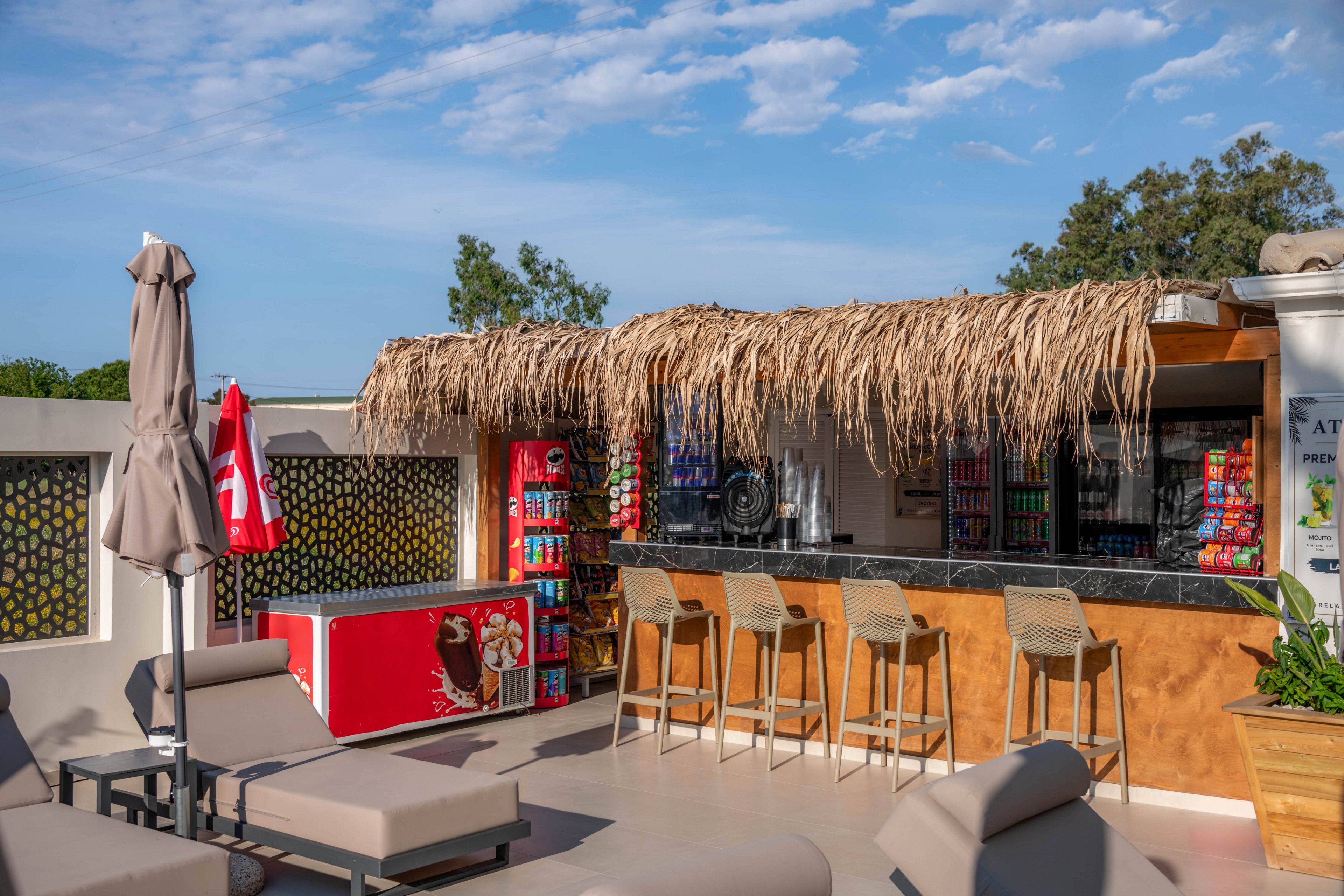 An outdoor tiki-style bar with a thatched roof stands beside a poolside lounge area. Several bar stools line the counter, while cushioned sun loungers and closed umbrellas sit in the foreground. Refrigerated drink coolers and colorful beverage displays are visible behind the bar. The scene is lit by bright daylight under a blue sky with scattered clouds, creating a relaxed vacation atmosphere.