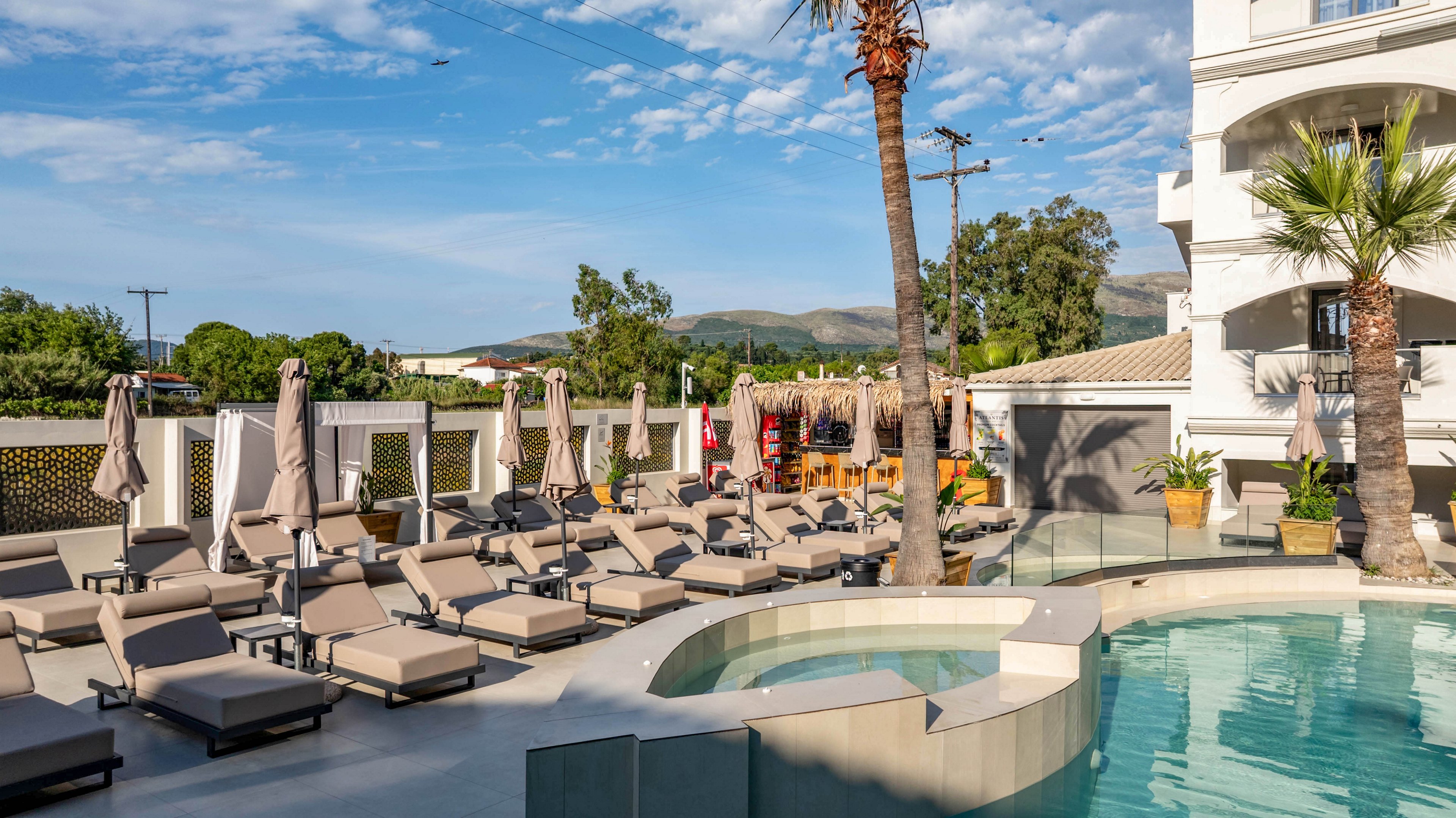 A contemporary outdoor hotel pool area is lined with numerous cushioned sun loungers and closed umbrellas. The curved pool features a shallow ledge and smooth tiled edges, reflecting the clear blue sky. Palm trees and surrounding greenery add a resort feel, while white buildings and distant hills frame the background. The scene suggests a quiet, sunny day at a vacation destination.