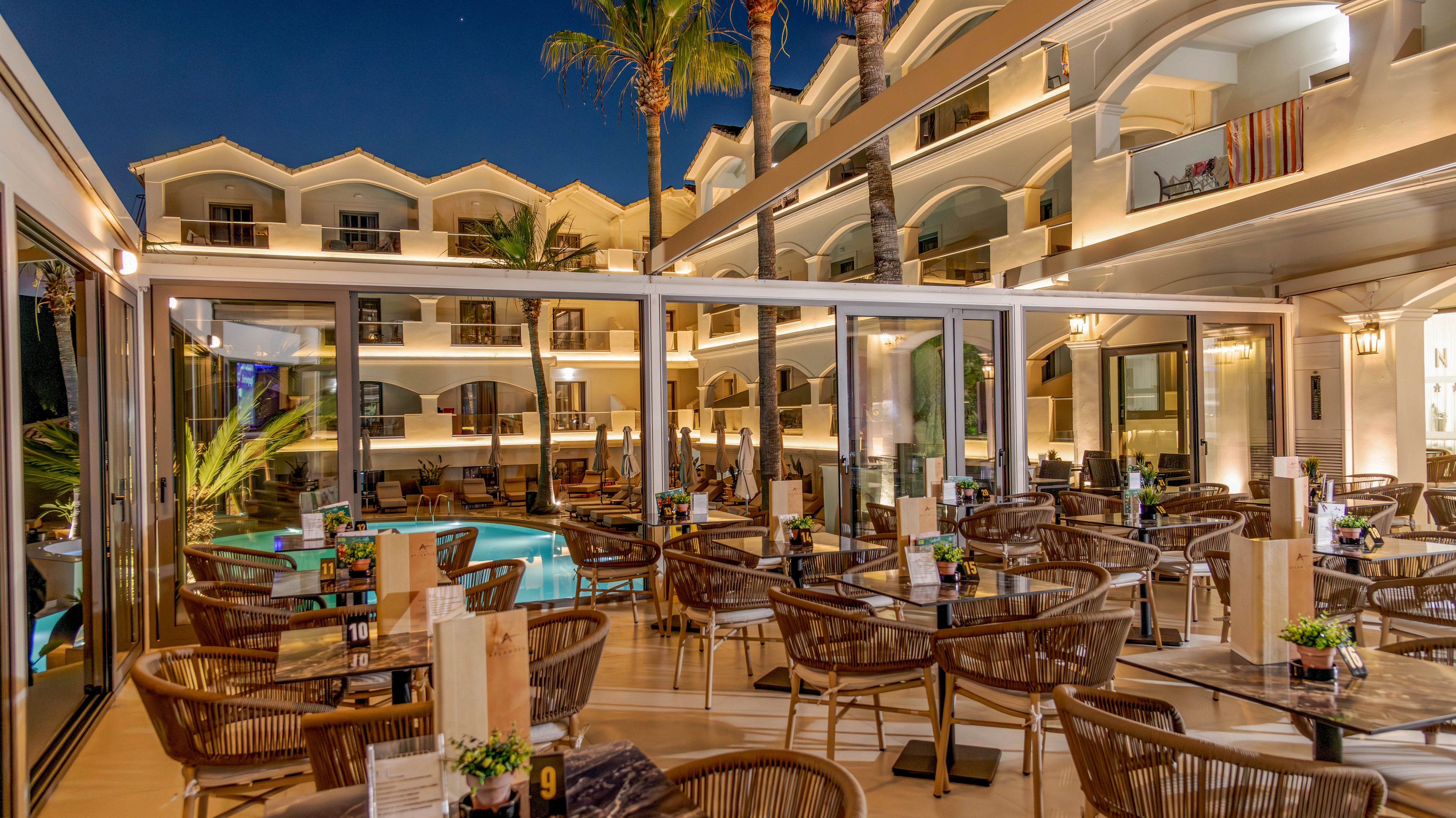 An elegant outdoor restaurant terrace is set beside a hotel swimming pool at night. Numerous wicker chairs and wooden tables are neatly arranged, each decorated with small flower arrangements and table numbers. Warm ambient lighting from the building and pool creates a cozy, inviting atmosphere under a clear evening sky. The surrounding multi-story hotel features balconies and palm trees, enhancing the resort-like setting.