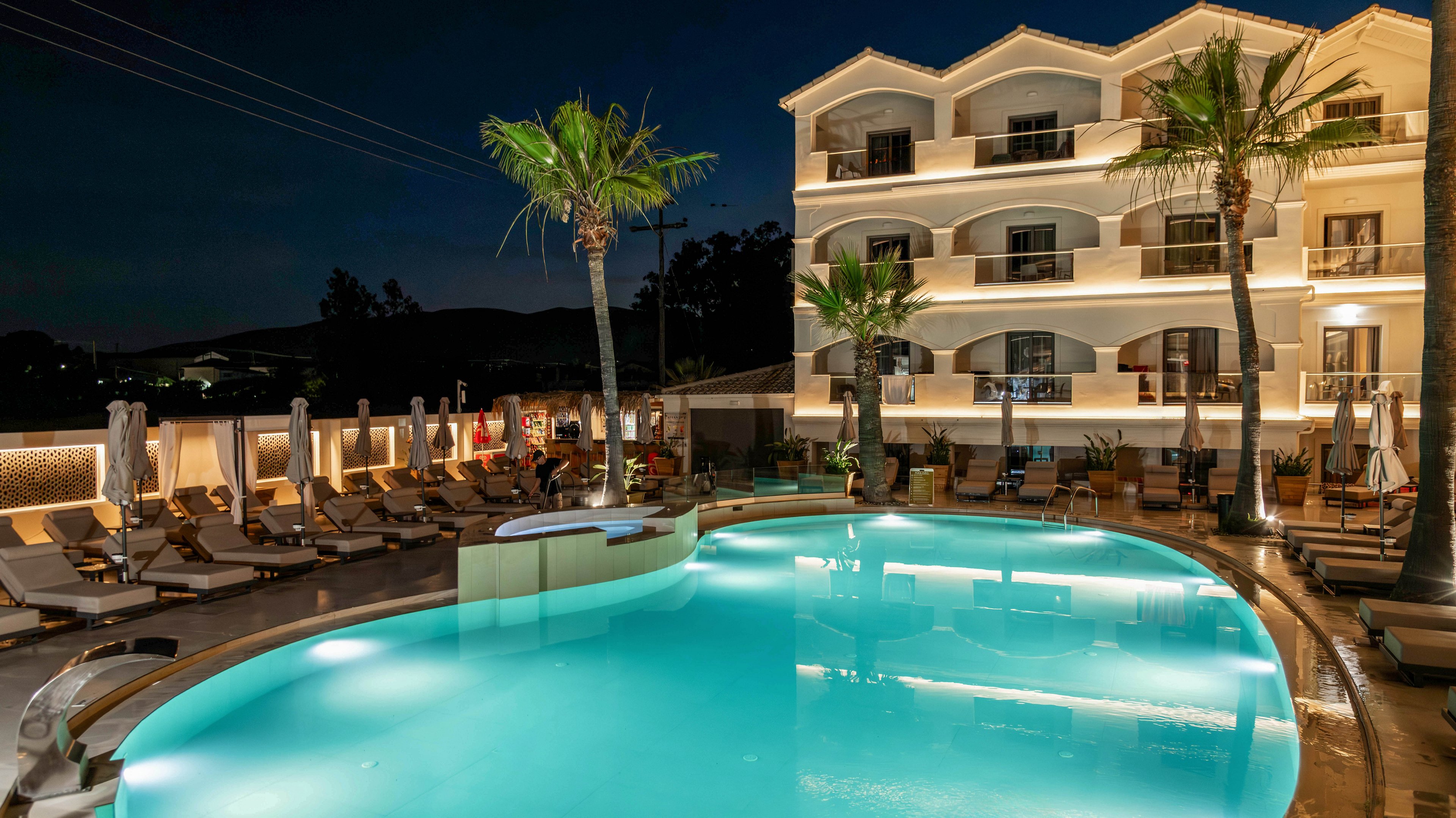 A modern multi-story hotel is shown at night with warm lights glowing from balconies and windows. In the foreground, a large curved swimming pool glows with turquoise water, reflecting the building and palm trees. Sun loungers and closed umbrellas line the poolside, suggesting a resort setting. The scene feels calm and inviting, emphasizing nighttime relaxation and vacation ambiance.