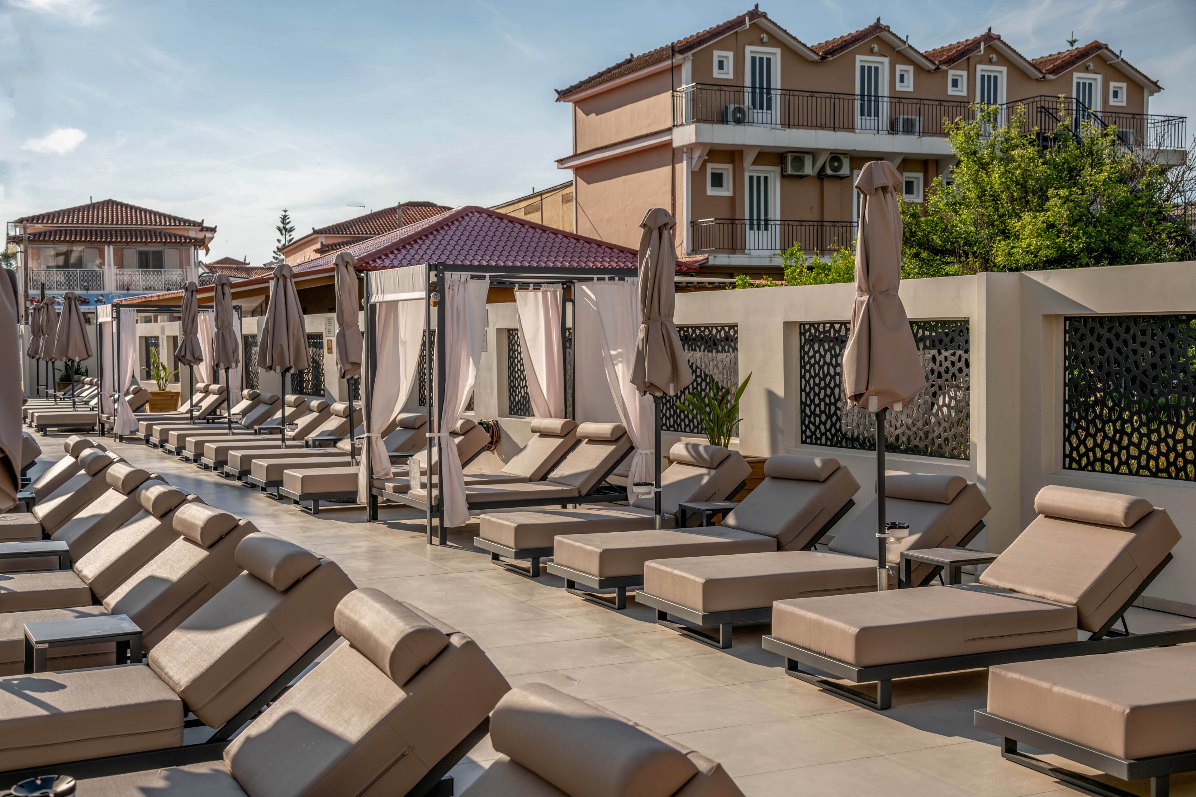A row of cushioned sun loungers lines a resort terrace, each paired with a closed umbrella. The scene appears calm and empty, suggesting early morning or off-peak hours at the pool area. In the background, multi-story hotel buildings with balconies overlook the deck. Warm neutral tones and soft lighting create a relaxed vacation atmosphere.