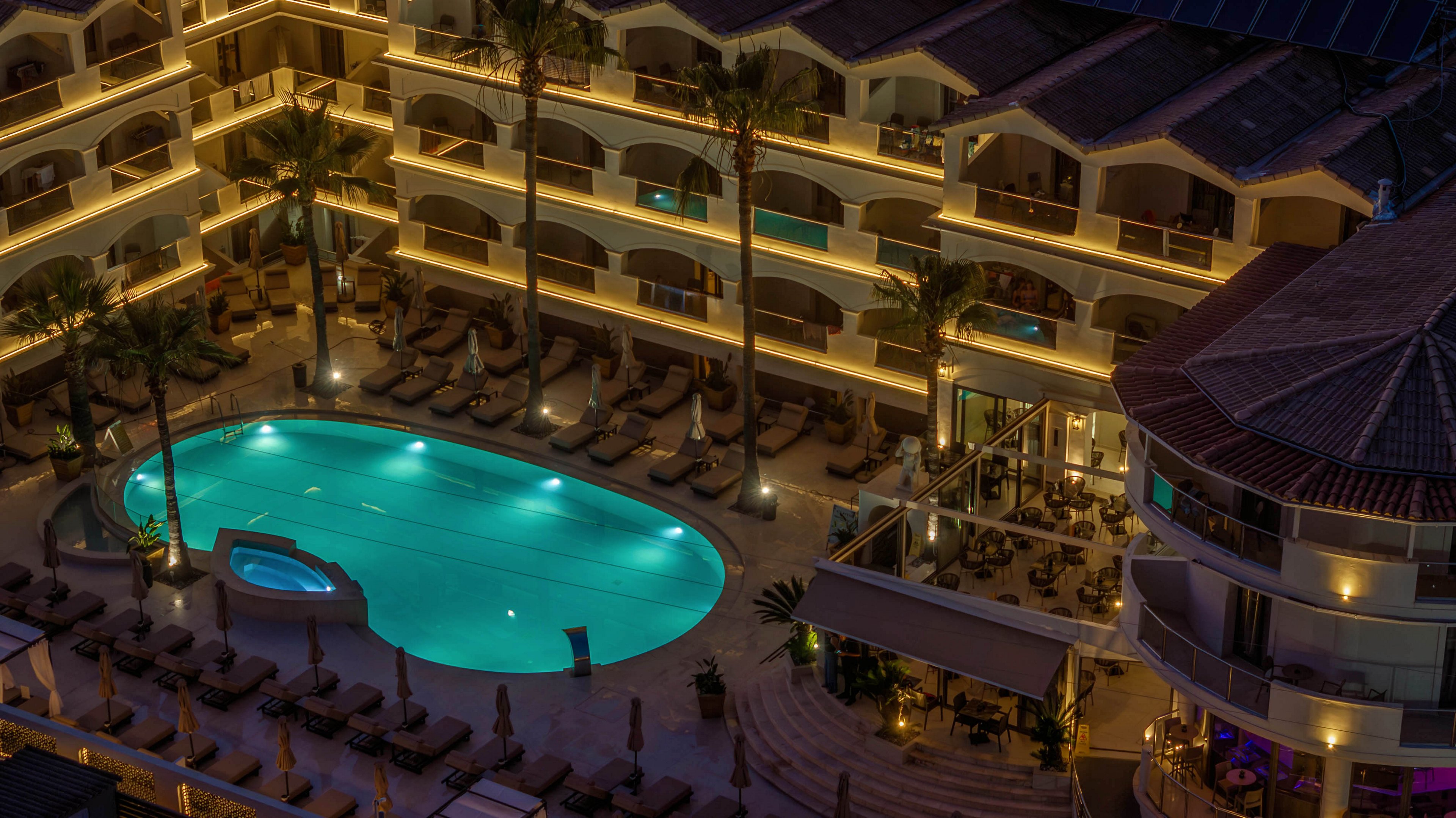 A large outdoor swimming pool glows with turquoise light at the center of a resort complex at night. Multi-story hotel buildings with balconies and warm accent lighting surround the pool area. Numerous sun loungers and palm trees line the deck, creating a relaxed vacation atmosphere. The scene suggests a tranquil evening setting at a coastal or tropical holiday destination.