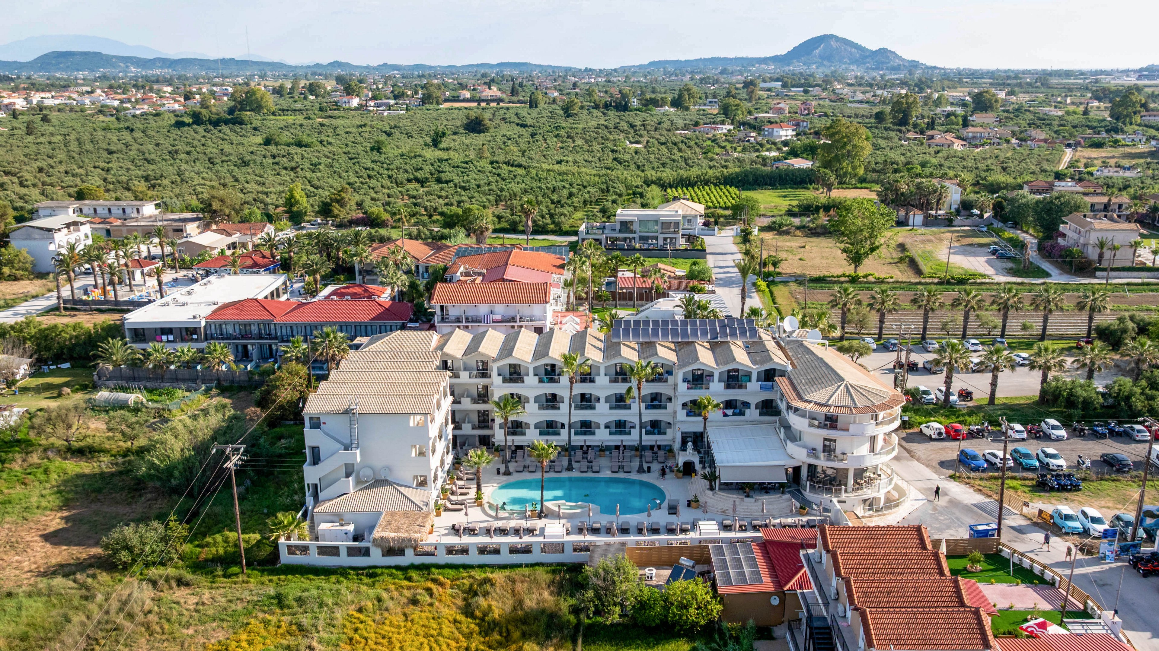 A large resort-style hotel complex with a central outdoor swimming pool is captured from an aerial perspective. The property is surrounded by green fields, scattered houses, and distant hills under a clear sky. Palm trees line the entrance and parking area, and several vehicles are visible near the hotel. The scene conveys a sunny, relaxed vacation atmosphere in a semi-rural setting.