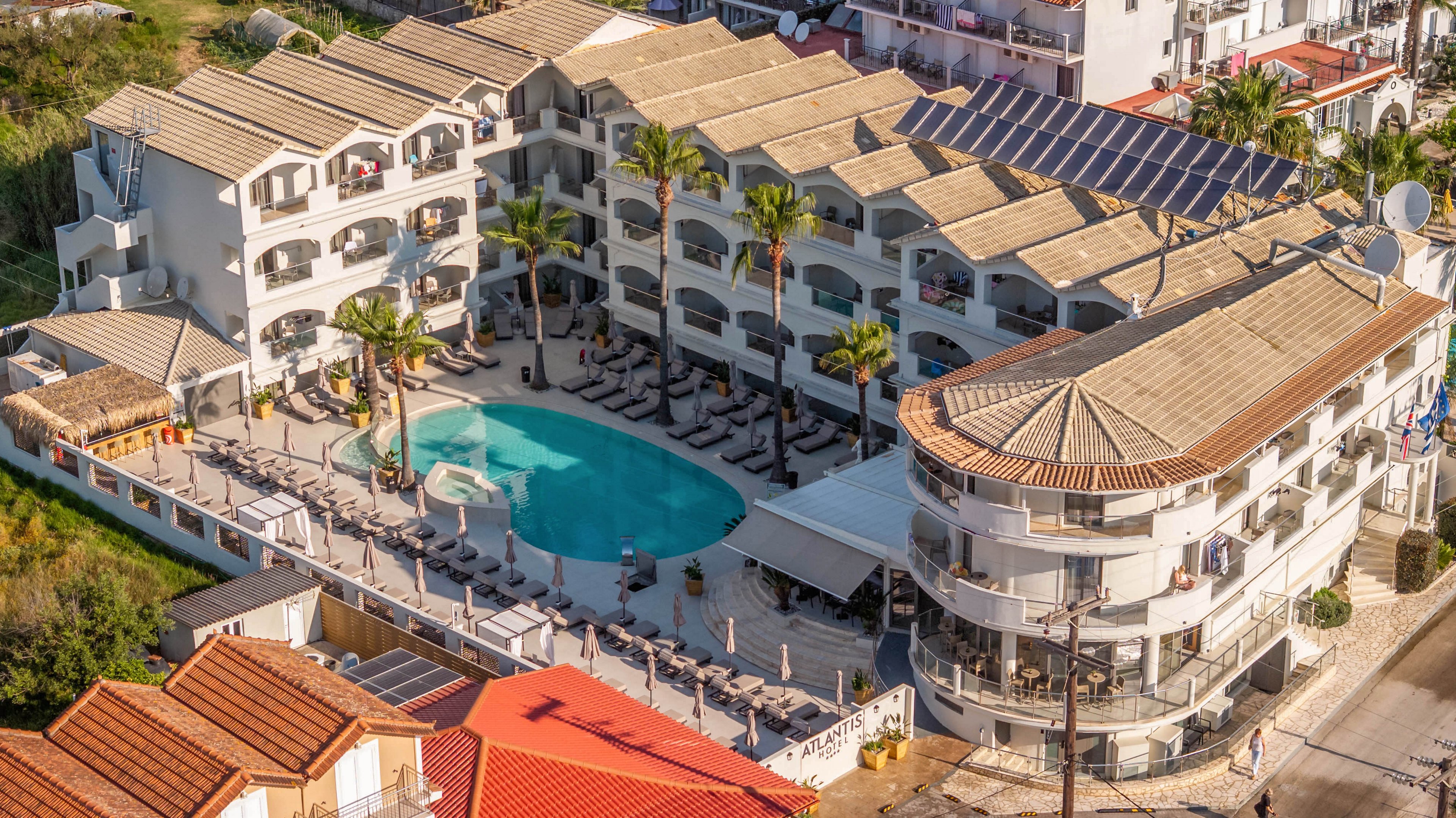 A large resort hotel complex is captured from above, showcasing a central turquoise swimming pool surrounded by sun loungers. The multi-story white buildings with tiled roofs form a courtyard around the pool, creating an enclosed, private atmosphere. Palm trees and shaded terrace areas add to the vacation setting, while nearby streets and neighboring houses indicate a coastal town location. The warm sunlight and clean architectural lines emphasize a relaxed, Mediterranean holiday mood.