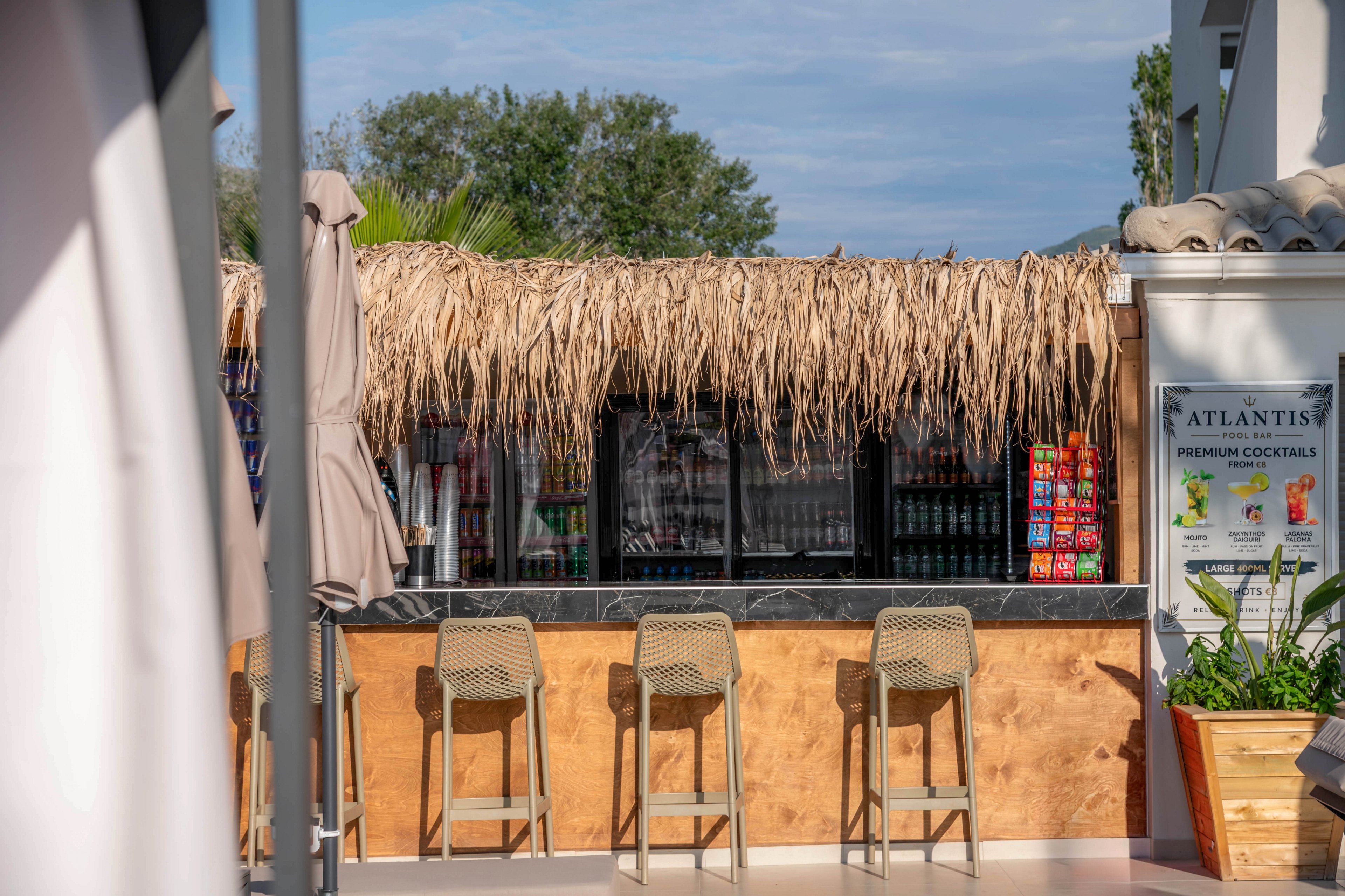 An outdoor tiki-style bar with a thatched roof stands empty under bright daylight. Four bar stools line the wooden counter, facing a shaded back bar stocked with drinks and snacks. Large umbrellas and nearby greenery suggest a relaxed resort or poolside setting. A sign on the right advertises premium cocktails at a venue named Atlantis.