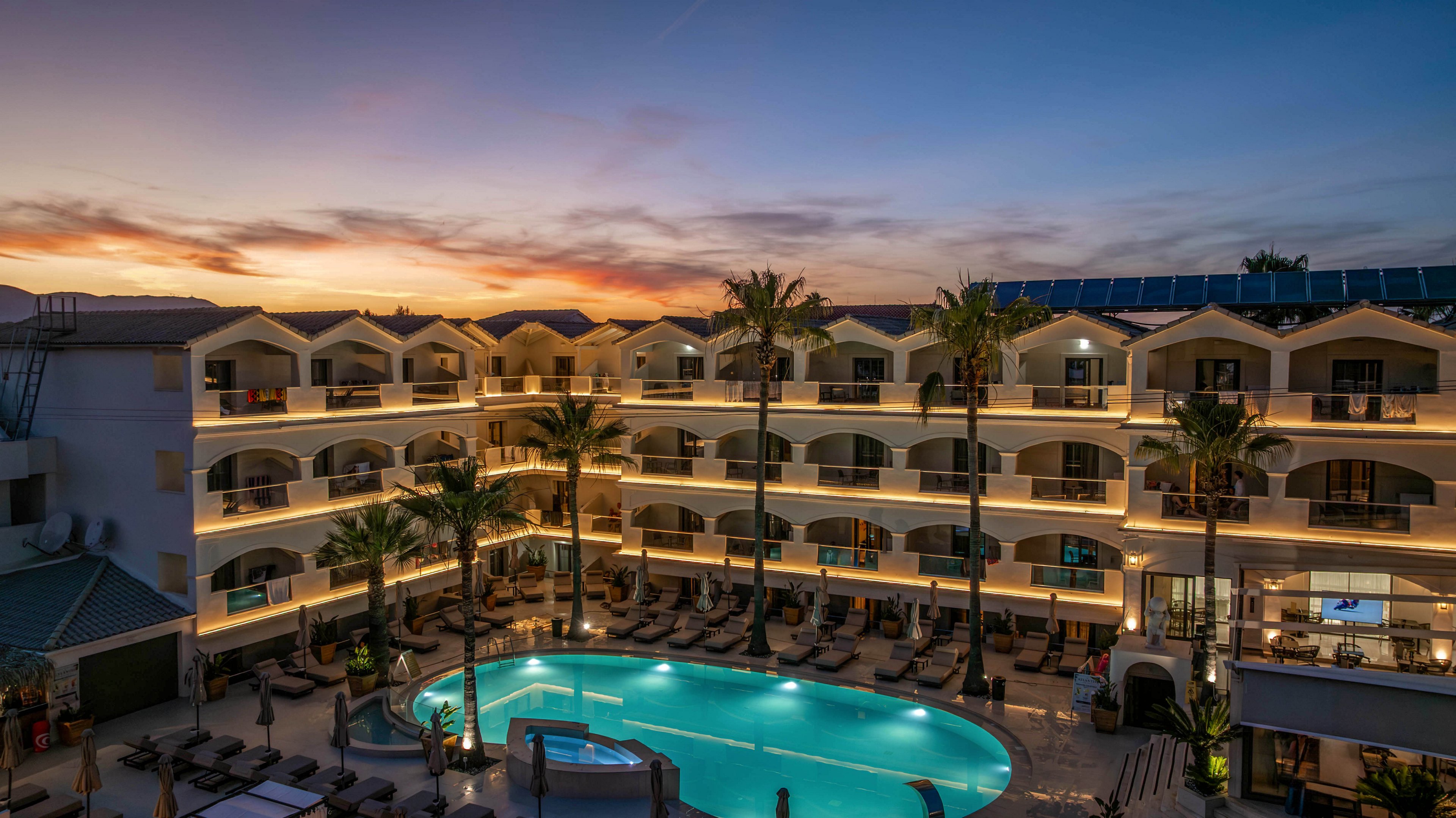 A multi-story resort hotel surrounds a large illuminated swimming pool at dusk. Warm lights outline the balconies and architecture, contrasting with the deep blue sky and subtle sunset colors. Palm trees frame the pool area, where lounge chairs are neatly arranged around the water. The tranquil atmosphere suggests a relaxing vacation destination in a warm climate.