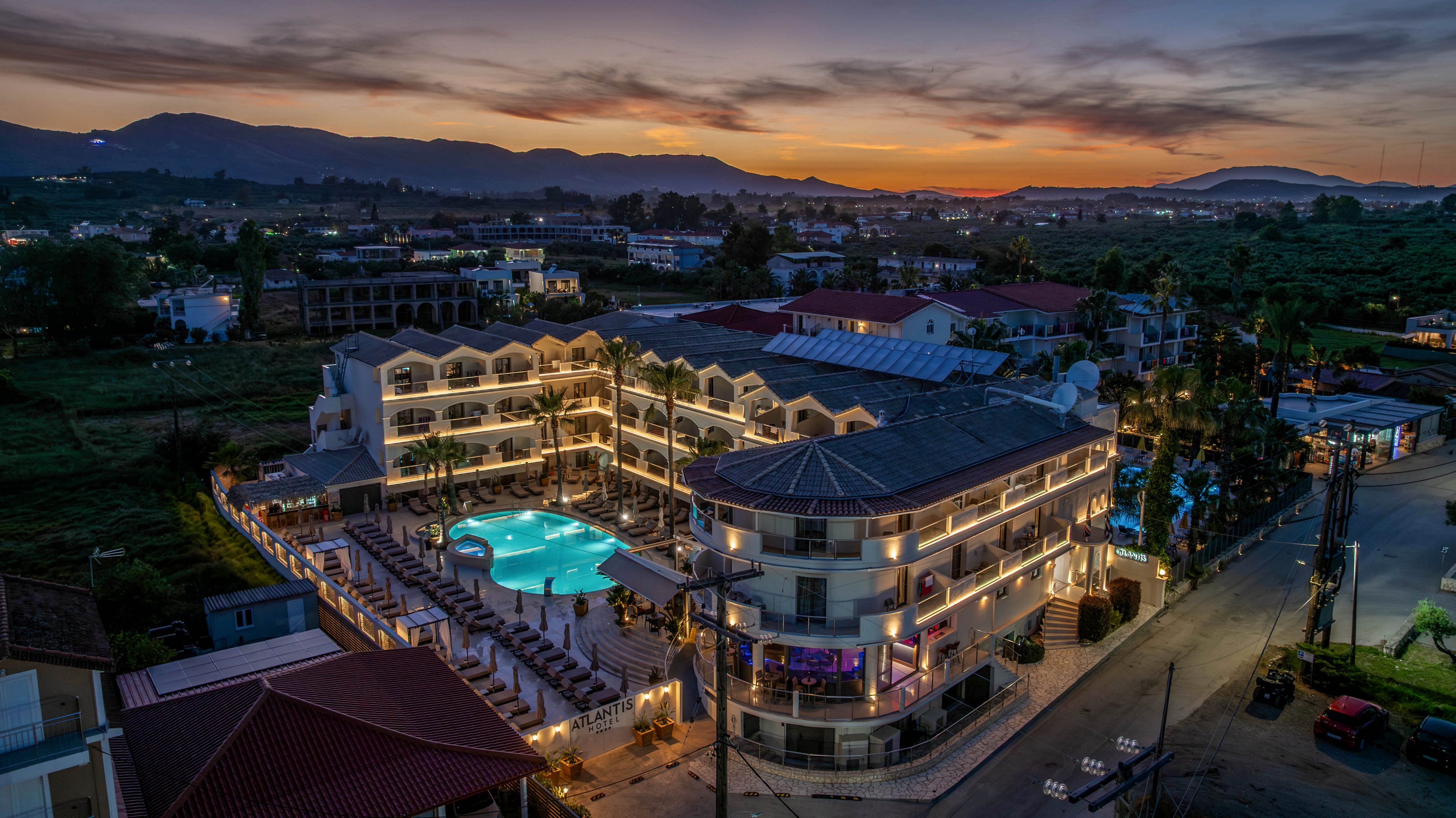 A modern resort complex is captured from above at twilight, with buildings wrapped around a glowing outdoor swimming pool. The surrounding landscape fades into fields and distant mountains under a colorful evening sky. Streetlights and building lights create reflections and highlights that emphasize the resort’s architecture. The scene conveys a tranquil vacation atmosphere as night begins to fall.