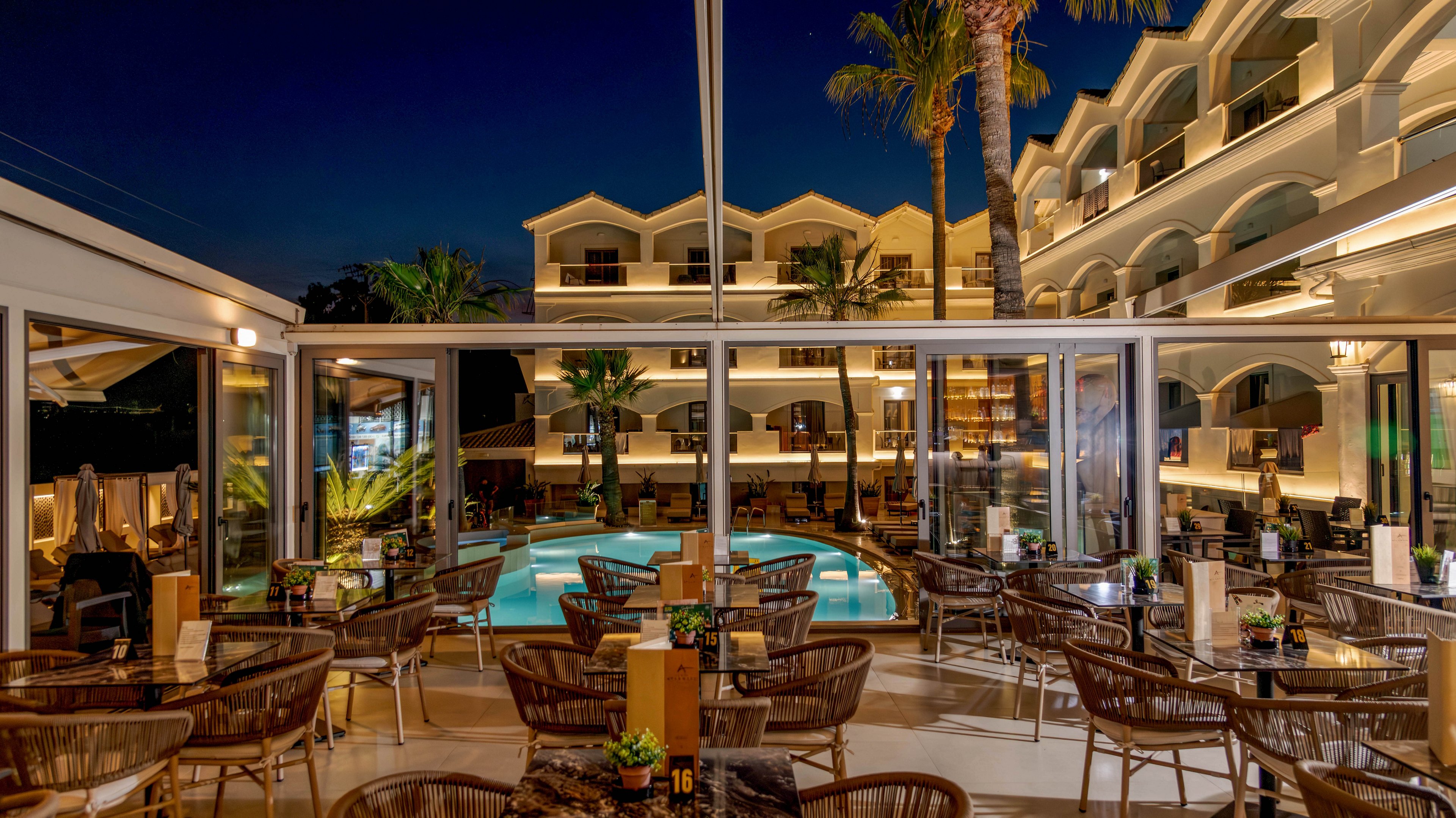 An elegant hotel restaurant overlooks a brightly lit swimming pool at night. Numerous wicker chairs and tables are arranged neatly on a covered terrace, each set with menus and table numbers. The surrounding hotel building features balconies and warm ambient lighting, creating a welcoming atmosphere. Palm trees and reflections in the pool enhance the luxurious vacation setting.