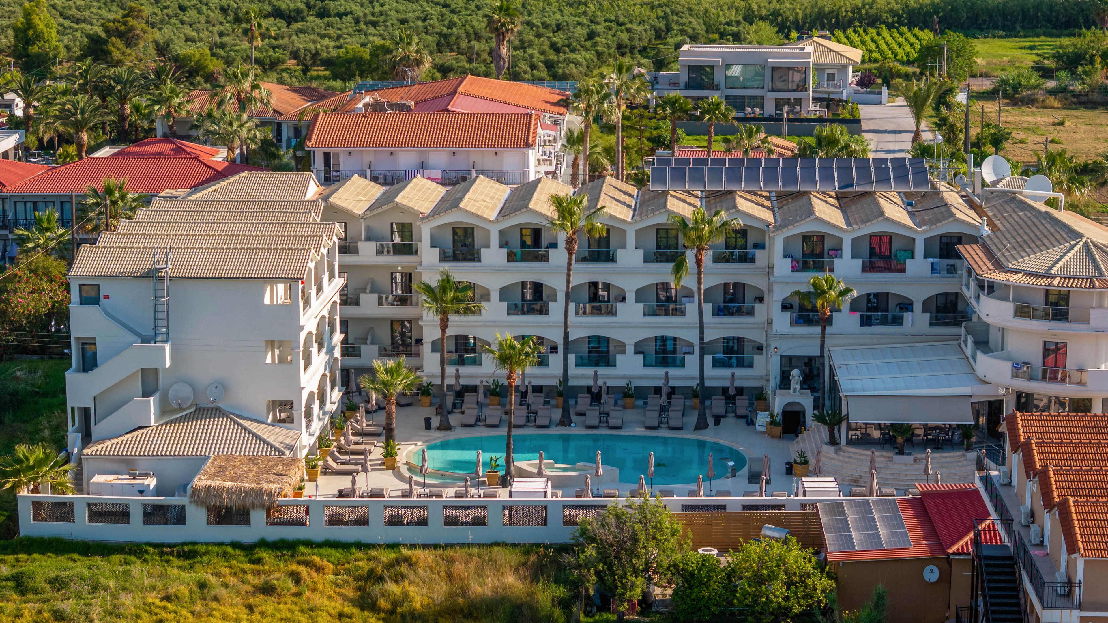 A multi-story resort hotel with a central outdoor pool is captured from an elevated aerial perspective. The building features rows of balconies facing the pool area, with sun loungers and umbrellas arranged around the water. Palm trees and landscaped greenery surround the complex, adding to the tropical atmosphere. In the background, additional buildings and cultivated fields extend toward the horizon under clear daylight.