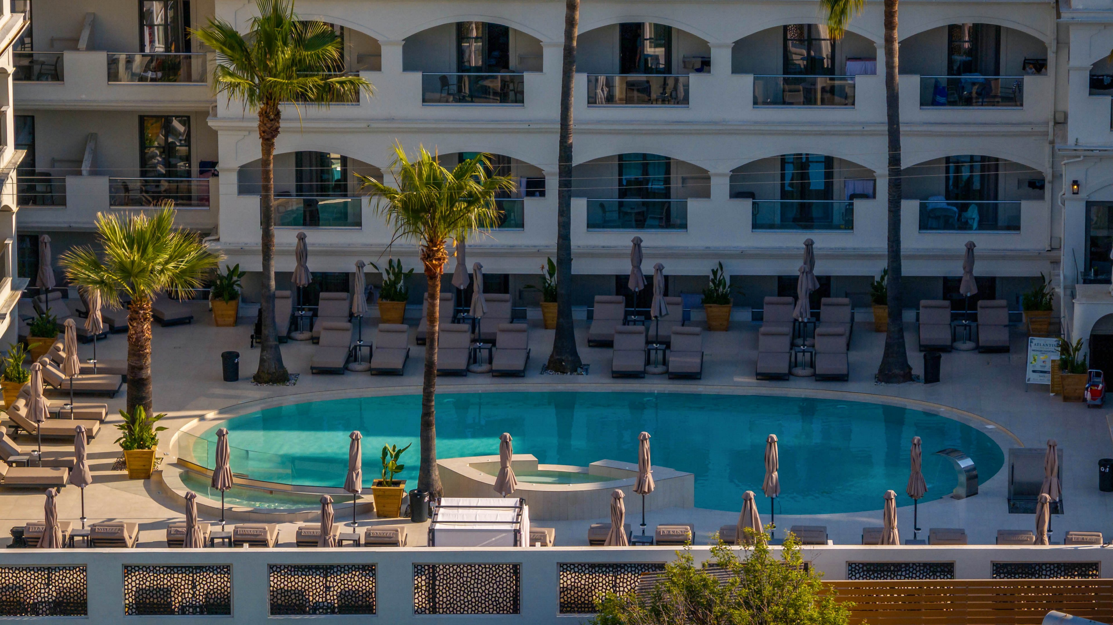A modern hotel courtyard features a circular outdoor swimming pool surrounded by rows of lounge chairs and umbrellas. Tall palm trees add a tropical touch to the clean, white multi-story building in the background. The balconies and arched windows overlook the pool area, suggesting a resort-style accommodation. Warm sunlight and clear blue water create a relaxed vacation atmosphere.
