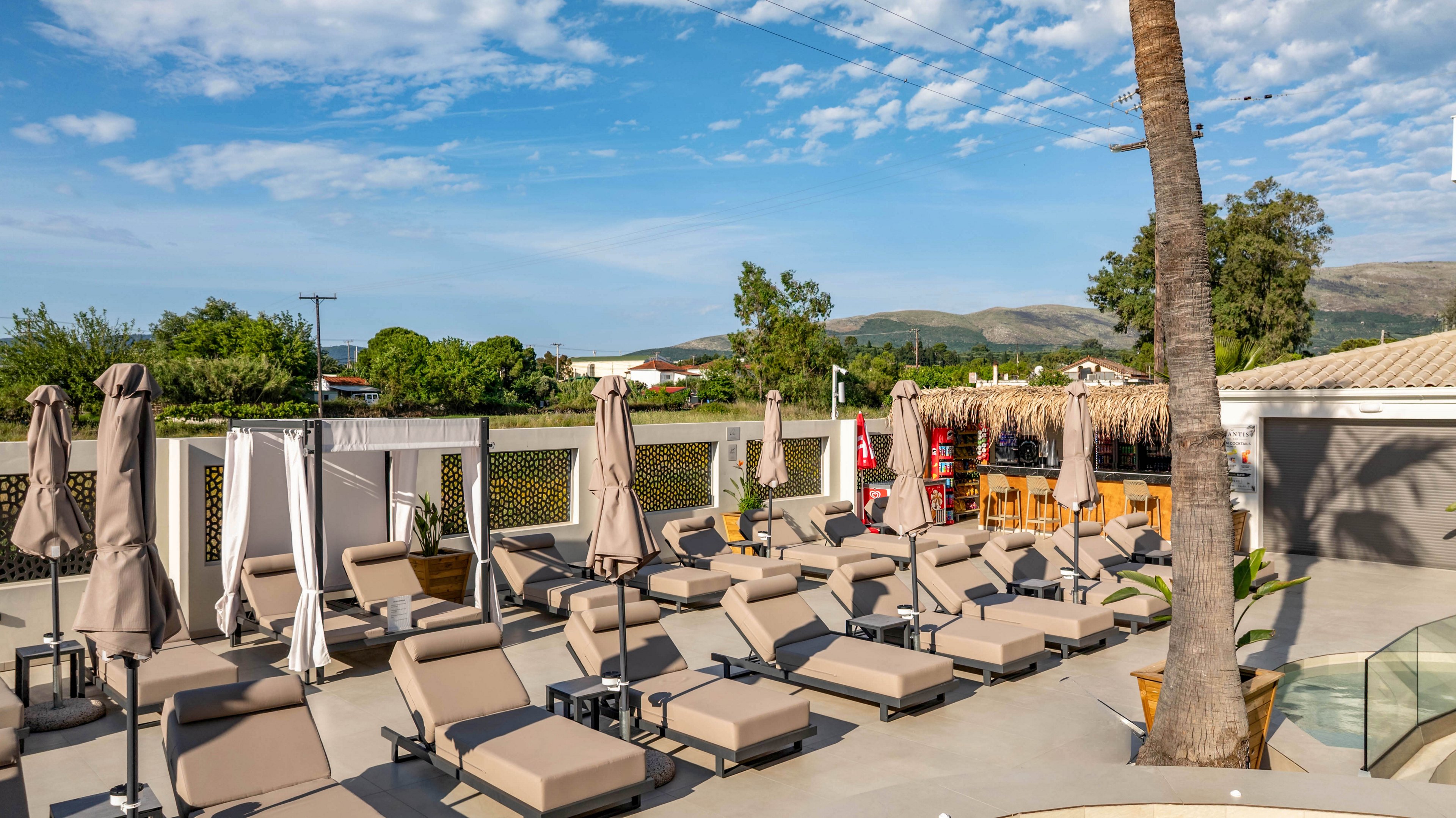 A modern outdoor resort terrace is lined with rows of beige cushioned sunbeds and closed umbrellas. The area appears quiet and empty, suggesting early morning or off-peak hours. A palm tree, wooden bar structure, and privacy fencing frame the lounging space. In the background, green trees, low buildings, and distant hills sit under a bright blue sky with scattered clouds.