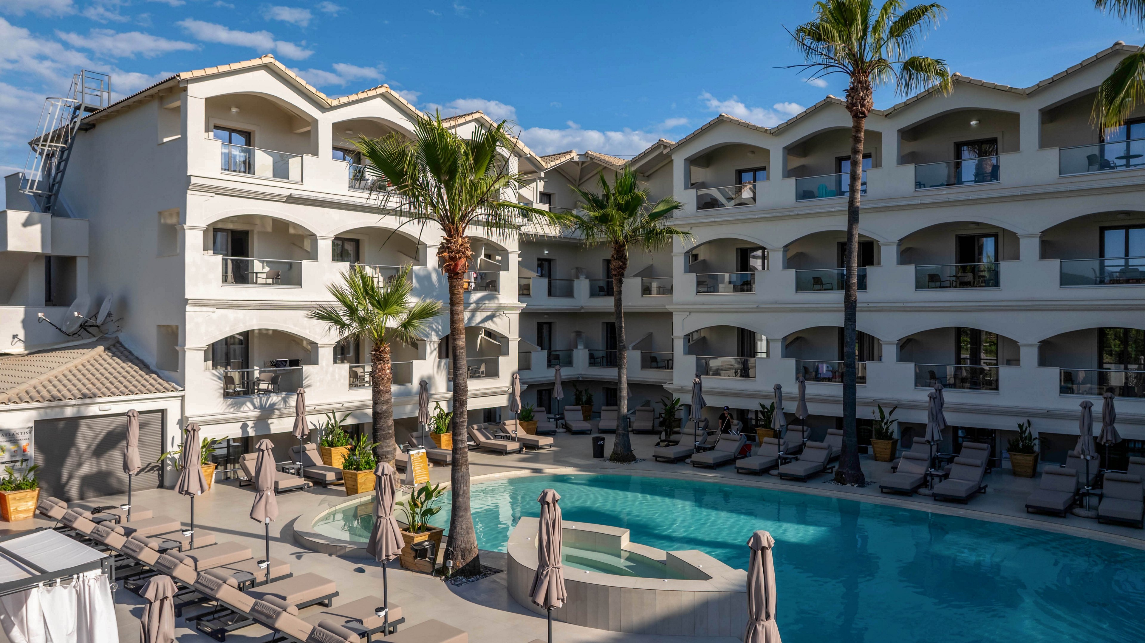A contemporary multi-story hotel surrounds a curved outdoor swimming pool. Sun loungers and closed umbrellas line the poolside terrace, suggesting a relaxing resort atmosphere. Tall palm trees rise between the balconies, adding a tropical touch to the white building facade. Clear blue sky and bright daylight emphasize a warm, inviting vacation setting.