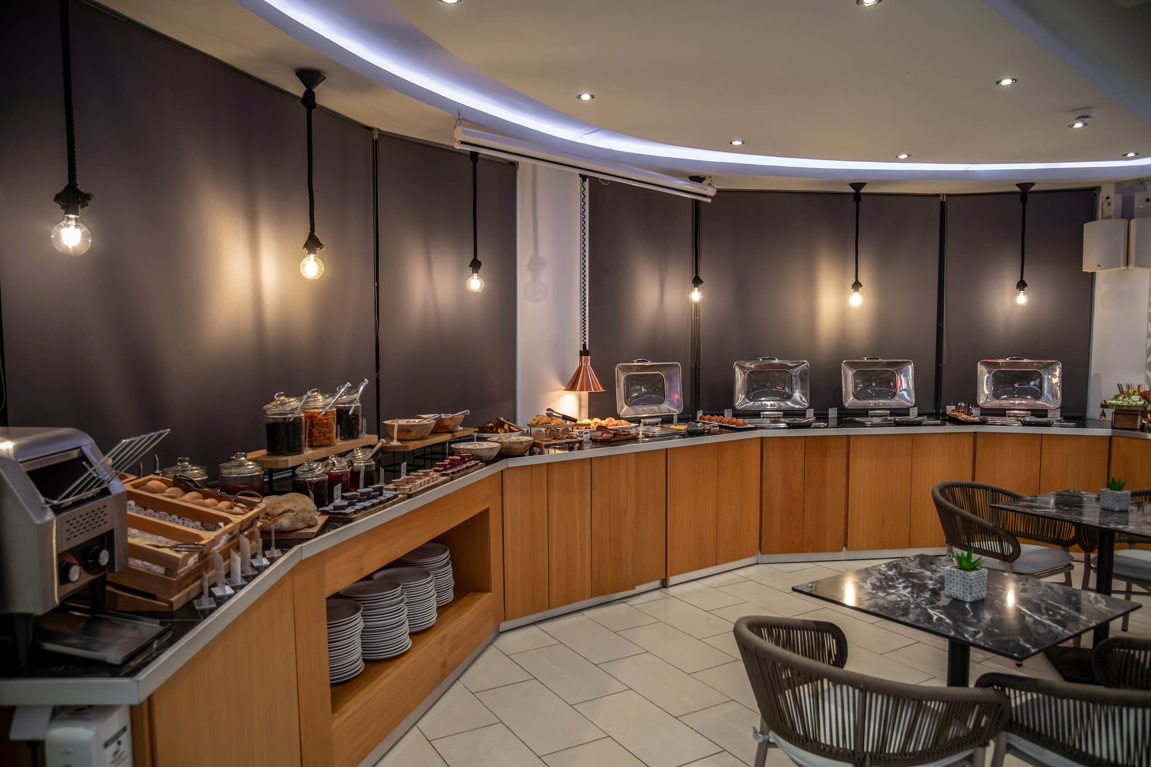 A modern self-service buffet is arranged along a curved wooden counter in a hotel dining area. Chafing dishes, cereal dispensers, bread trays, and condiments are neatly organized for guests. Warm pendant lights hang from the ceiling, casting a cozy glow against dark wall panels. Empty chairs and marble-topped tables in the foreground suggest the space is ready for diners.