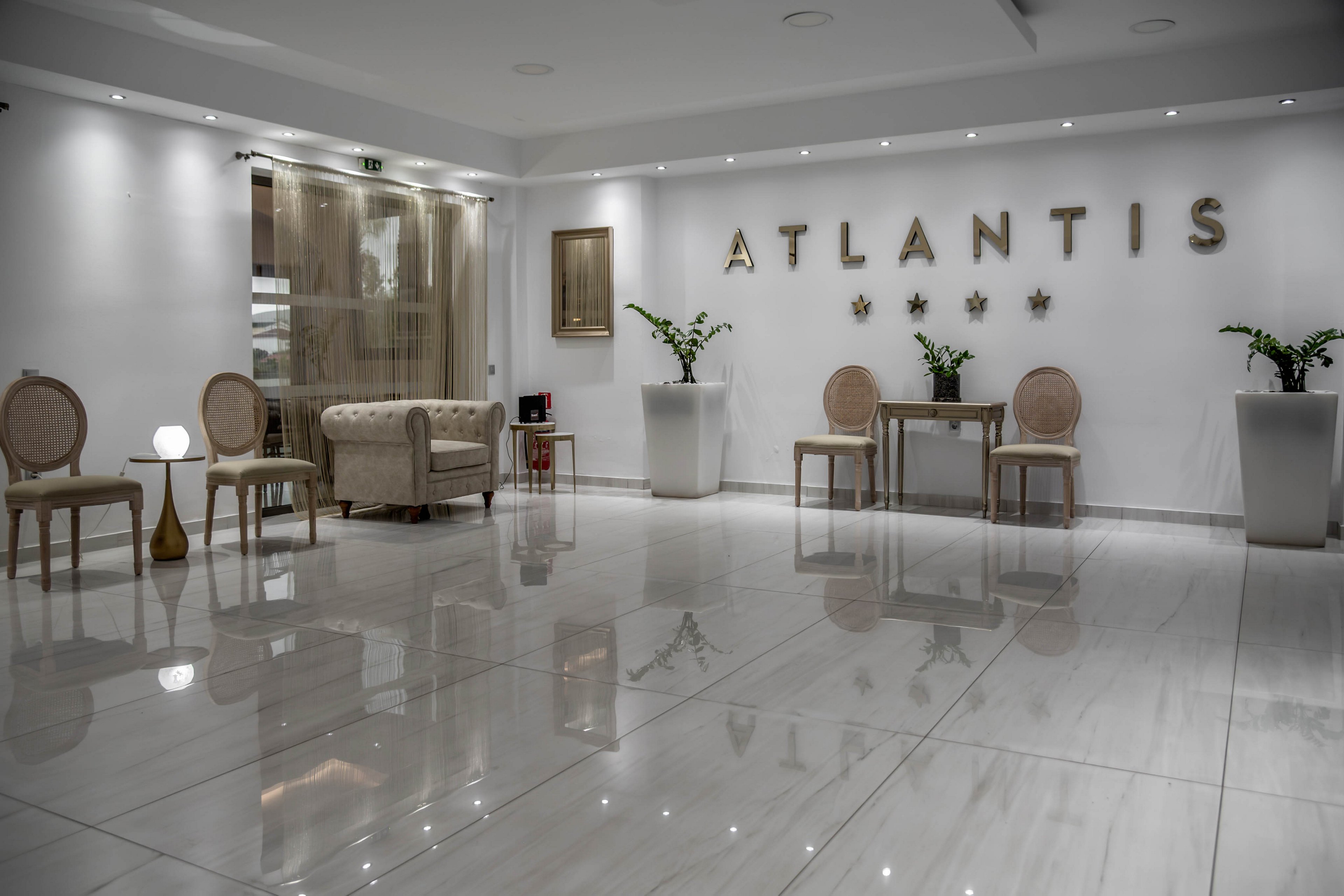 Spacious modern hotel lobby featuring the word ATLANTIS and five stars on the wall. The room has glossy marble floors that reflect the furniture and lighting. Elegant chairs, a small sofa, and side tables are arranged along the walls with decorative plants in tall white planters. Soft, neutral colors and subtle spotlights create a calm, upscale atmosphere.