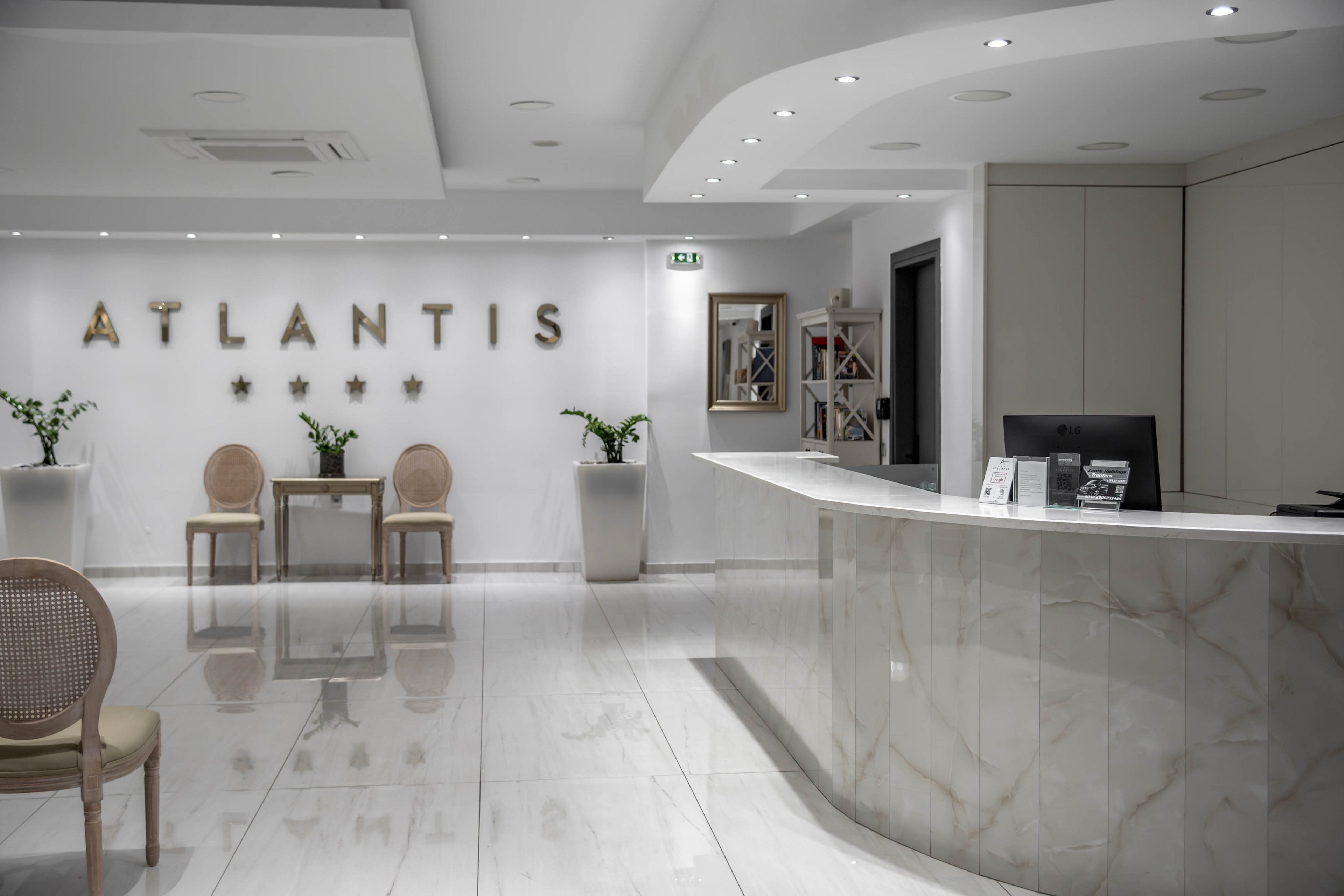 Bright, contemporary hotel reception area featuring a curved marble front desk. The white lobby has glossy tiled floors and recessed ceiling lights creating a clean, minimalist look. The word "ATLANTIS" and four stars are displayed on the back wall above two chairs and a small table with plants. A bookshelf, wall mirror, and potted plant add subtle decorative accents to the calm, welcoming space.