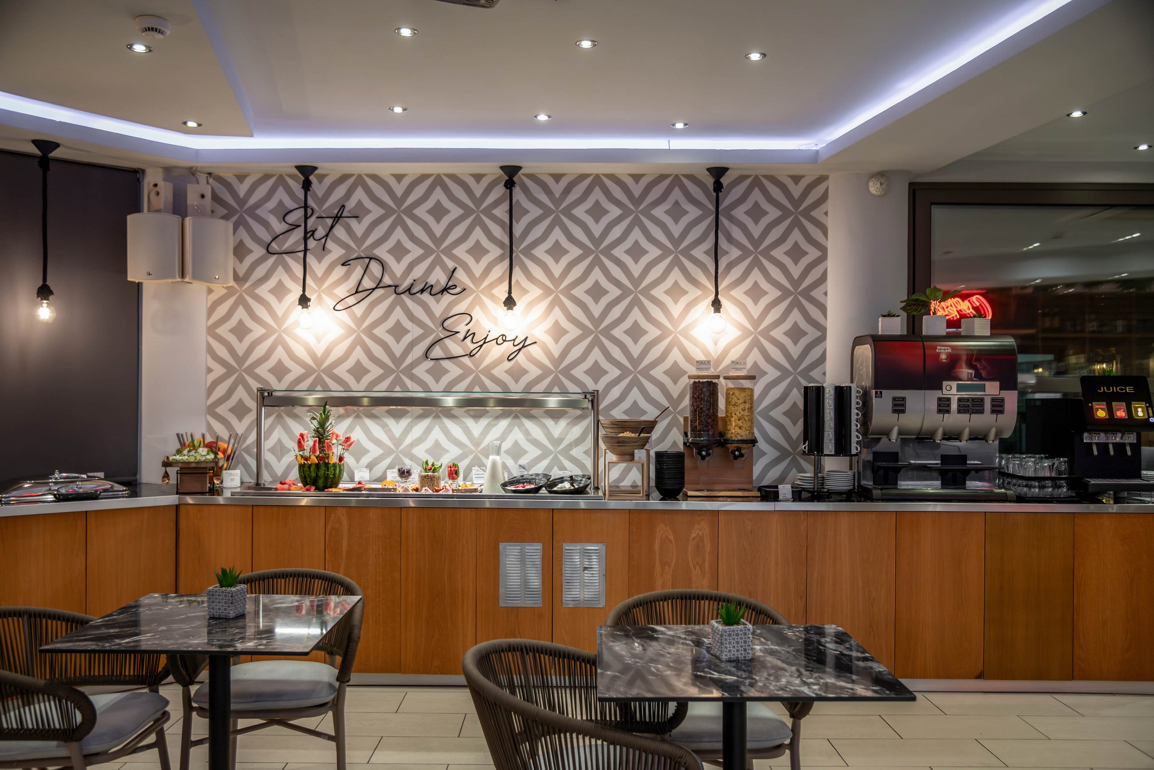 Contemporary self-service buffet area in a hotel or café with wooden counters and food warmers. The background wall features geometric patterned tiles and decorative script text reading "Eat Drink Enjoy." A coffee machine, juice dispensers, and neatly arranged condiments line the countertop. In the foreground, marble-top tables with wicker chairs create a relaxed dining space under recessed ceiling lights.