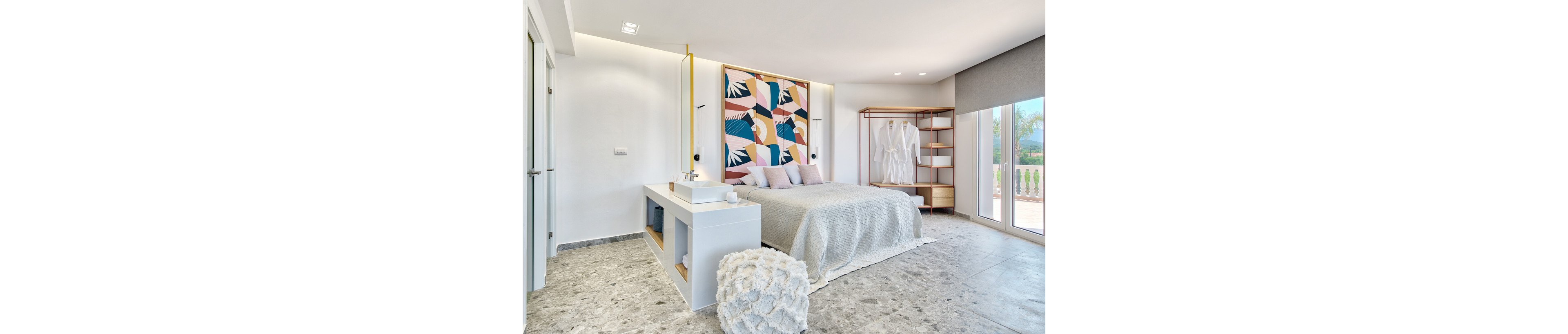 A contemporary bedroom features a large bed with a textured light gray cover and multiple pillows. The room is accented by a bold, colorful abstract art piece above the headboard and an open wardrobe with white robes. Natural light fills the space through large windows, highlighting the minimalist decor and marble flooring.