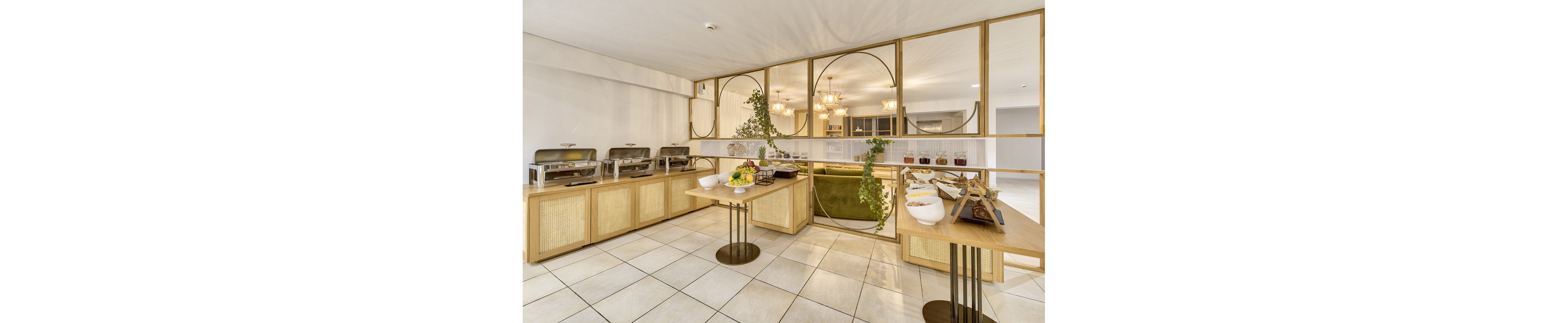 A contemporary buffet setup is displayed in a bright, spacious hotel dining area. The scene features wooden counters with stainless steel chafing dishes, bowls of fresh fruit, and baskets of breadsticks. Decorative greenery and geometric gold accents enhance the stylish ambiance, while warm lighting fixtures create an inviting atmosphere. The tiled floor and open layout contribute to a clean, modern look.