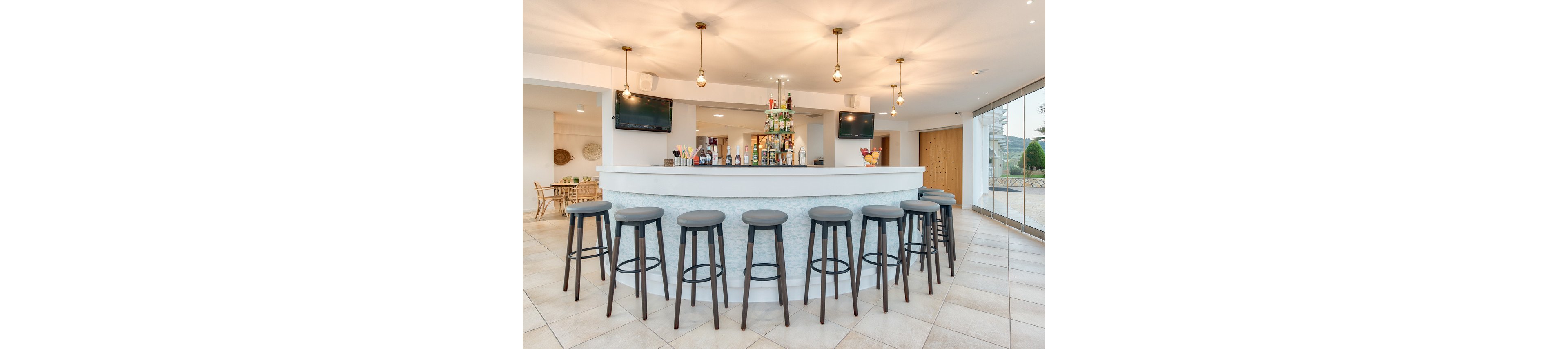 A contemporary hotel bar features a curved white counter surrounded by eight tall gray stools. The setting is bright and open, with pendant lights overhead and two wall-mounted televisions visible. The bar is stocked with various bottles and glassware, and large windows allow natural light to fill the space. The overall mood is clean, inviting, and modern, with neutral tones and minimalist decor.