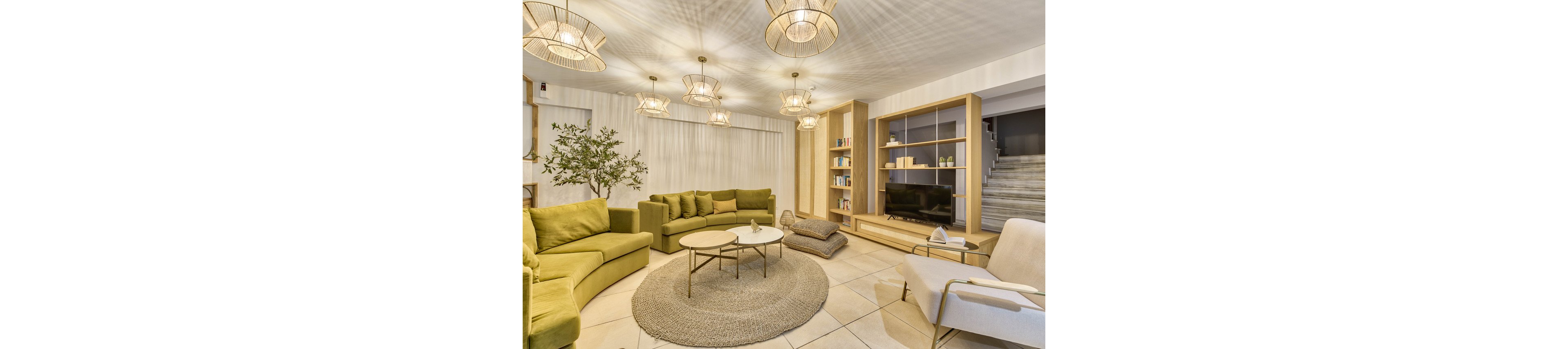 A spacious living room features green sectional sofas arranged around a pair of round coffee tables on a woven rug. The setting includes built-in wooden shelving with books and a television, soft overhead lighting, and a potted tree for a natural touch. Neutral tones and minimalist decor create a cozy, inviting atmosphere. The room also includes a staircase leading to an upper floor.