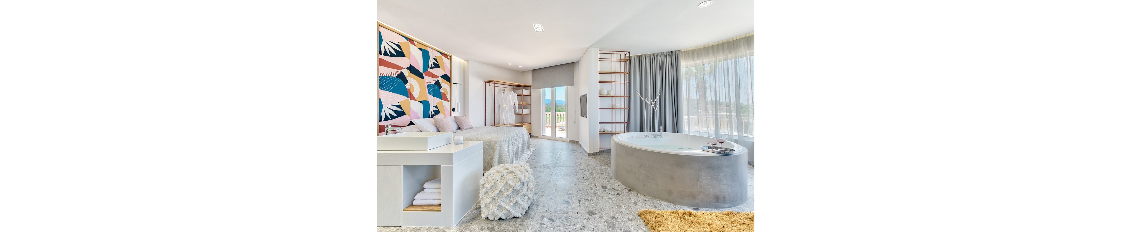 A spacious, contemporary bedroom features a large round bathtub and a bed with textured bedding. The room is decorated with a colorful geometric accent wall and minimalist wooden shelving. Natural light floods in through large windows with sheer curtains, and a balcony view is visible. The setting is serene and inviting, with soft neutral tones and pops of color.