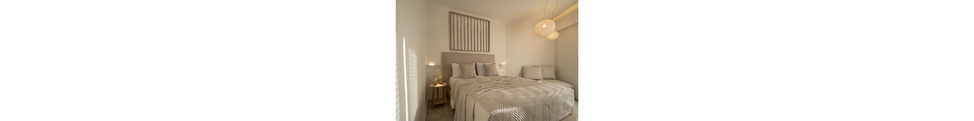 A contemporary bedroom features a large bed with textured beige bedding and multiple pillows. The setting is minimalistic, with soft, warm lighting from woven pendant lamps and wall sconces. A small side table holds a decorative item, and a cushioned bench sits at the foot of the bed. The overall palette is neutral, with beige and cream tones creating a calm, inviting atmosphere.