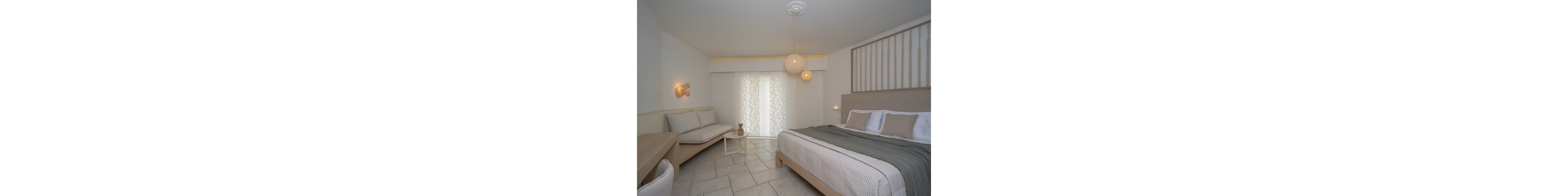 A contemporary hotel room features a large bed with neutral bedding and accent pillows. The space includes a cozy seating area, a wooden desk, and pendant lighting, all set against a backdrop of soft beige and white tones. Sheer curtains filter natural light, creating a calm and inviting atmosphere. The room is uncluttered, with clean lines and a minimalist aesthetic.