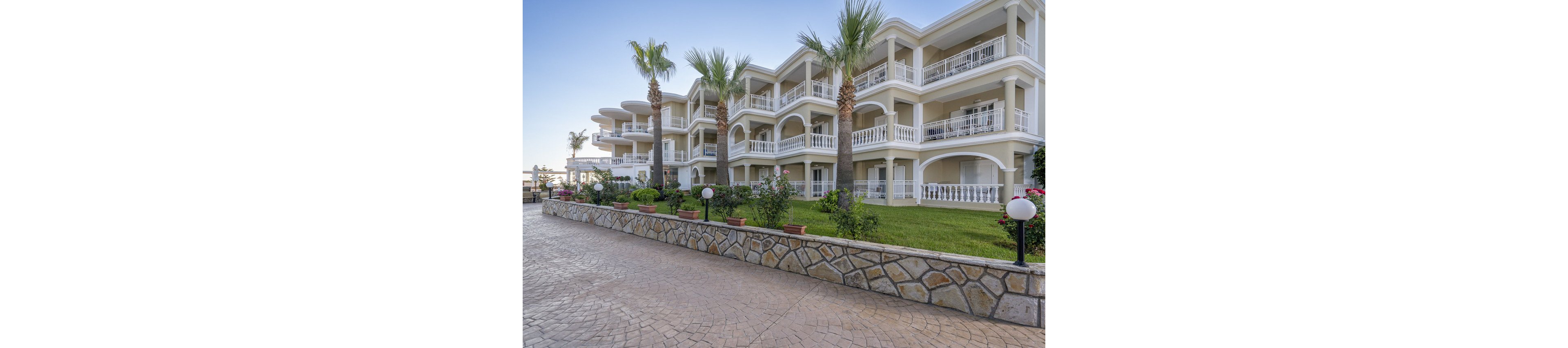 A multi-story hotel building features white railings, spacious balconies, and Mediterranean architectural elements. The setting includes manicured gardens, palm trees, and a paved walkway in the foreground. The scene is captured in daylight with a clear blue sky, evoking a calm and inviting atmosphere. No visible text or numbers are present in the image.