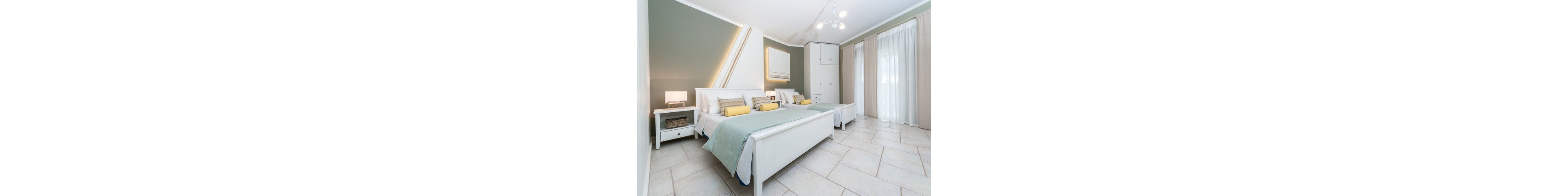 A contemporary bedroom features two neatly made beds with pastel green bedding and yellow accent pillows. The room is styled with clean white furniture, including nightstands and a wardrobe, and is illuminated by a modern chandelier and bedside lamps. Large windows with sheer curtains allow natural light to fill the space, creating a calm and inviting atmosphere. The overall palette is soft and neutral, enhancing the minimalist aesthetic.