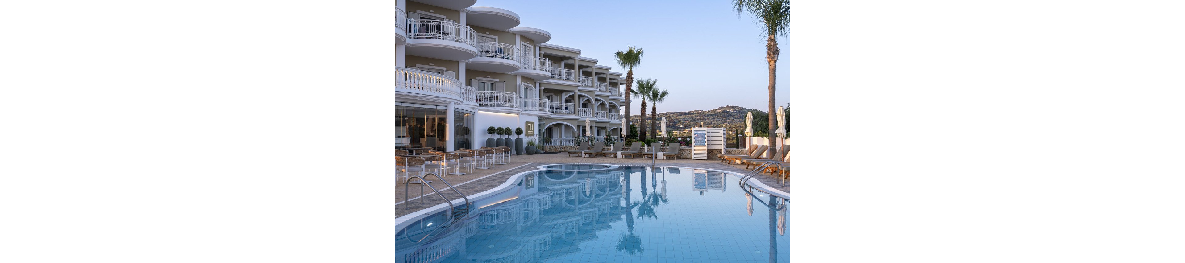 A contemporary multi-story hotel features spacious balconies overlooking a large, clear swimming pool. The setting is tranquil, with palm trees and lounge chairs arranged around the poolside, suggesting a relaxing vacation atmosphere. The architecture is elegant with white railings and soft neutral tones, and the surrounding landscape includes distant hills. No visible people or text are present in the image.