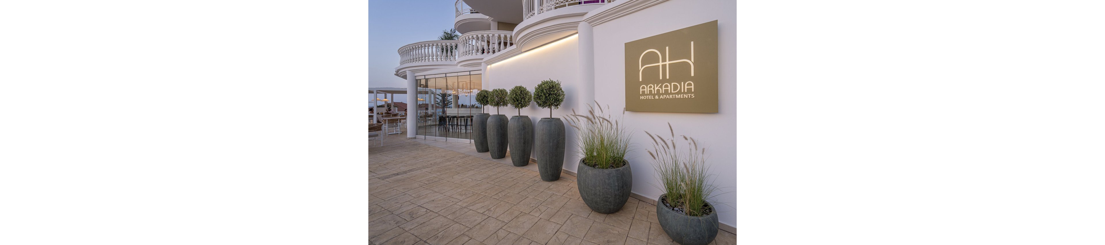 The image shows the entrance of Arkadia Hotel & Apartments, featuring a modern architectural design with curved balconies and large glass windows. Several tall, elegant planters with manicured trees and grasses line the tiled walkway, creating a welcoming atmosphere. The hotel sign with the text 'ARKADIA HOTEL & APARTMENTS' is clearly visible on the wall. The setting appears calm and inviting, with soft evening light enhancing the neutral color palette.