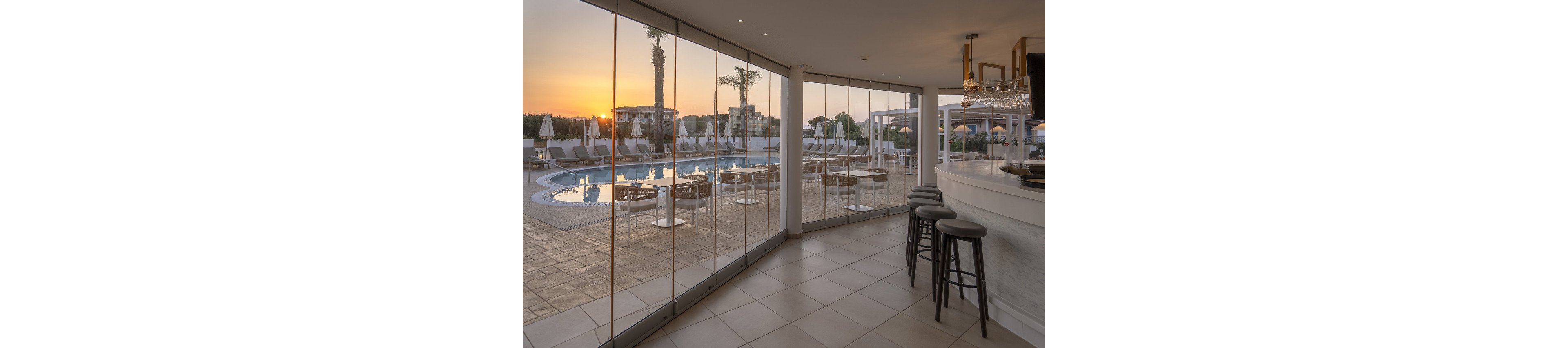 A contemporary bar with high stools overlooks a serene outdoor swimming pool through floor-to-ceiling glass windows. The scene is set at sunset, casting a warm glow over the pool area with lounge chairs and umbrellas. The interior features clean lines, neutral tones, and a relaxed atmosphere, while the outdoor setting highlights palm trees and resort buildings. No visible text or numbers are present in the image.