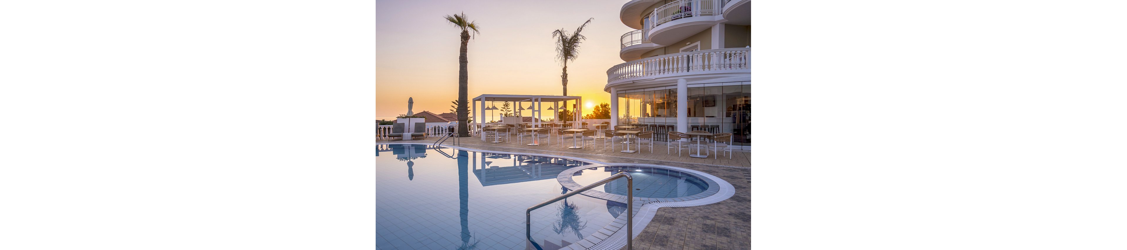 A modern hotel features a curved poolside terrace with empty tables and chairs, set against a tranquil sunset sky. Tall palm trees and a glassy swimming pool reflect the warm evening light, creating a serene and inviting atmosphere. The architecture includes white railings and rounded balconies, enhancing the upscale, resort-like ambiance. No people are present, emphasizing the peaceful mood. The scene is dominated by soft pastel and golden tones.