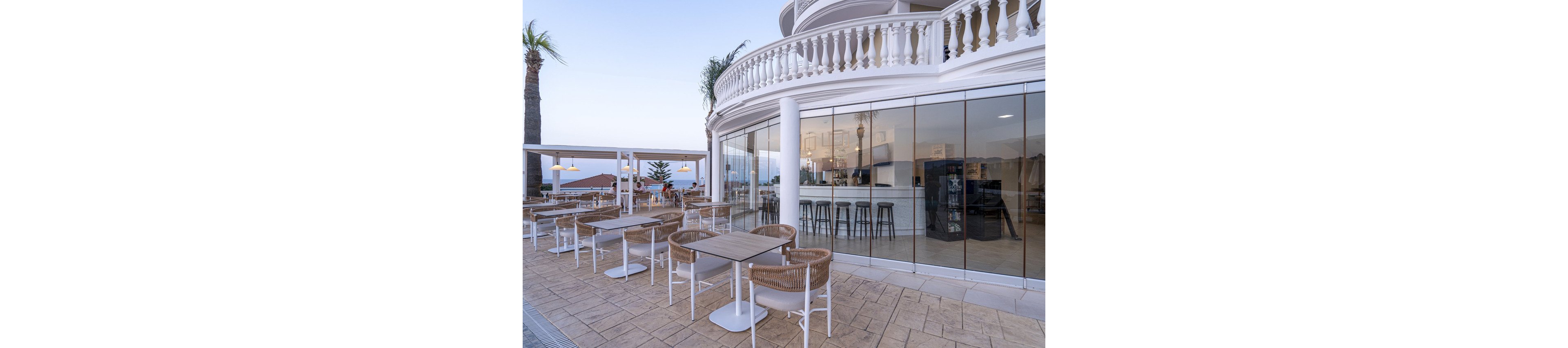 A contemporary outdoor cafe features wicker chairs and wooden tables arranged on a spacious tiled terrace. The setting includes a curved building with large glass windows, reflecting the bar area and stools inside. Palm trees and a clear sky create a relaxed, coastal atmosphere. The color palette is neutral with light browns, whites, and soft blues. No visible text or numbers are present in the image.