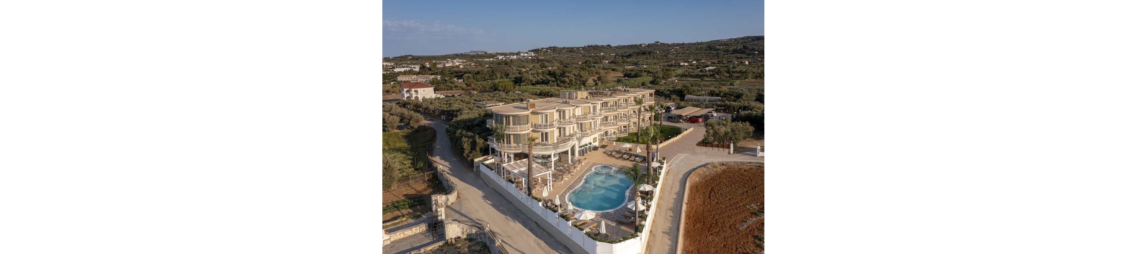 A contemporary multi-story hotel is captured from above, featuring a large outdoor swimming pool surrounded by sun loungers and umbrellas. The property is enclosed by a white fence and set in a rural landscape with fields and greenery. The building's curved balconies and clean architectural lines stand out against the natural backdrop. The scene is bathed in warm daylight, creating a welcoming and relaxed atmosphere.