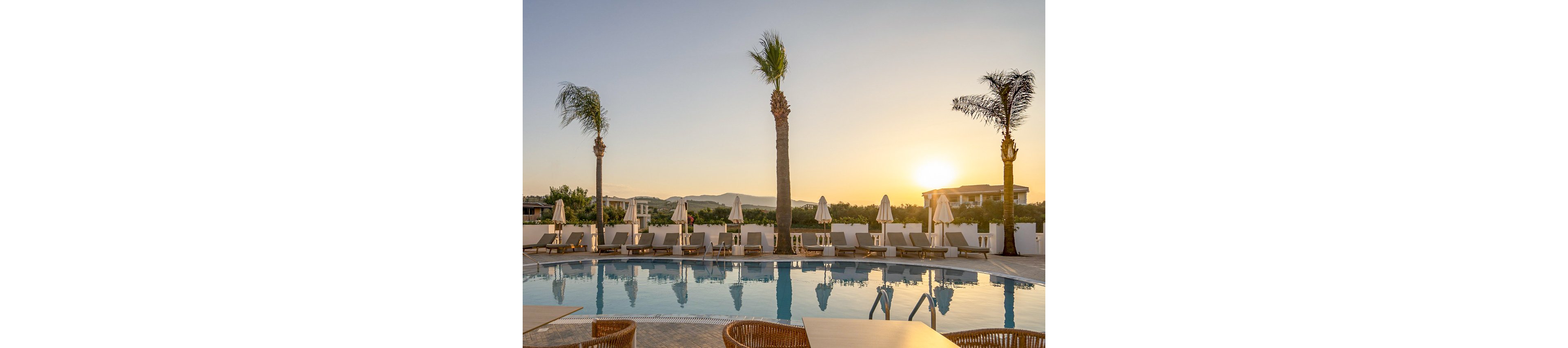 A tranquil resort pool area is captured at sunset, featuring tall palm trees and neatly arranged lounge chairs with umbrellas. The calm water reflects the golden sky, creating a peaceful and inviting atmosphere. Empty tables and chairs in the foreground suggest a relaxing, quiet moment before guests arrive. The setting is outdoors, with distant mountains visible in the background.